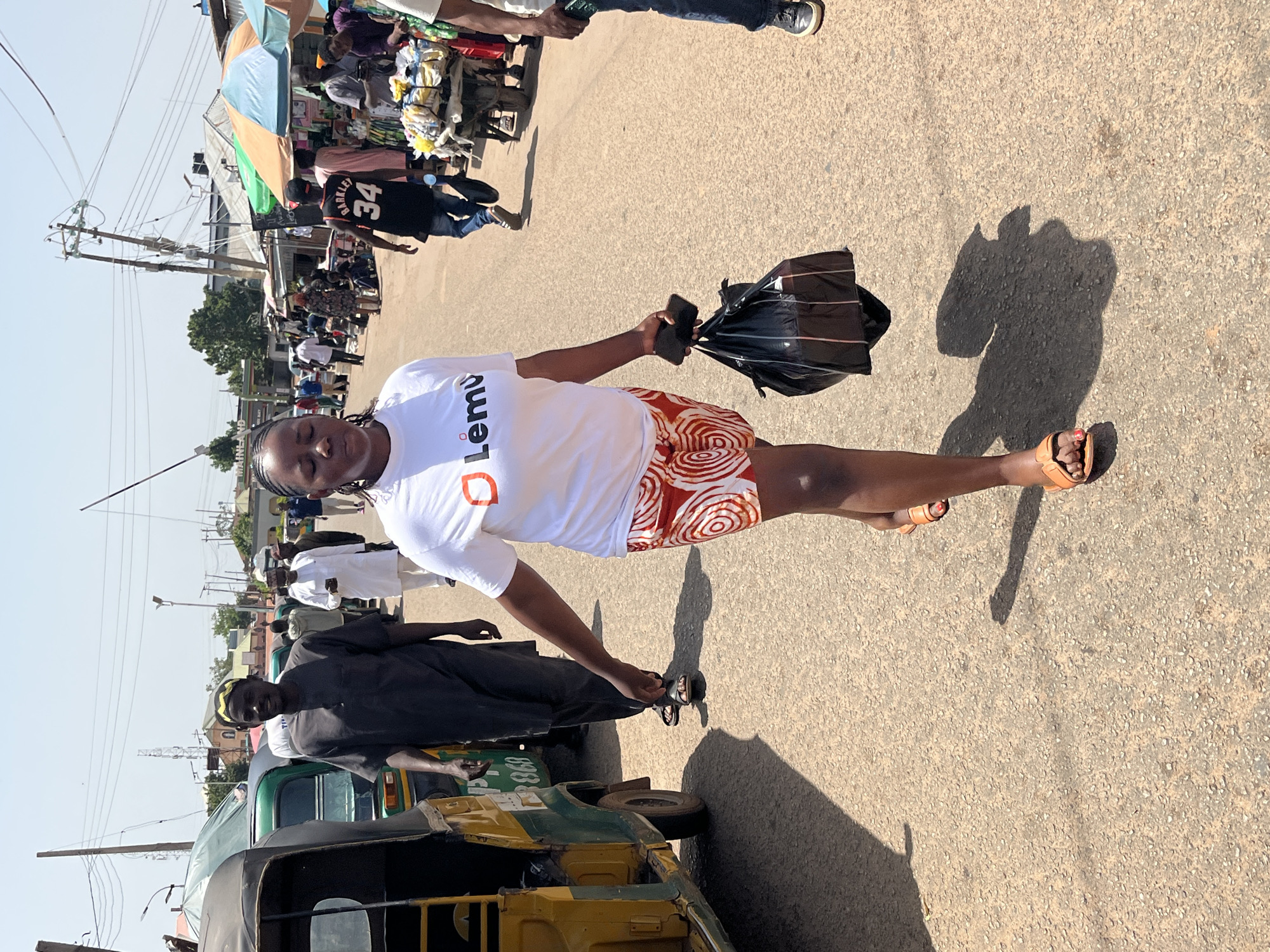 A medium shot shows a woman in the foreground, smiling at the camera. She is wearing a white t-shirt with an orange logo and a patterned skirt. She is holding a black plastic bag and a cell phone. Her feet are bare, wearing sandals. The background is a busy street with people, stalls, and a three-wheeled vehicle. The scene appears to be outdoors during the day, with bright sunlight casting shadows.
There is visible text on the woman's t-shirt, which appears to be "Lėmù" and an orange symbol. The three-wheeled vehicle has a license plate with the number "0938". The overall atmosphere seems lively and bustling.