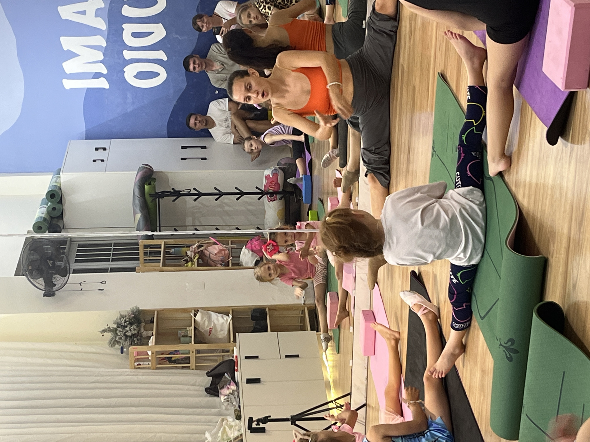 In a yoga studio in Bac Nha Trang Ward, Vietnam, an instructor in an orange tank top and grey pants demonstrates a split pose on a green yoga mat. Several young girls are also on yoga mats, some attempting similar poses. Two young girls in pink are seated on the floor, facing the instructor. In the background, adults are seated, observing the yoga session. The studio features large mirrors reflecting the scene, shelves with various items, and a mural on the wall. The floor is wooden, and yoga mats are arranged for the participants.