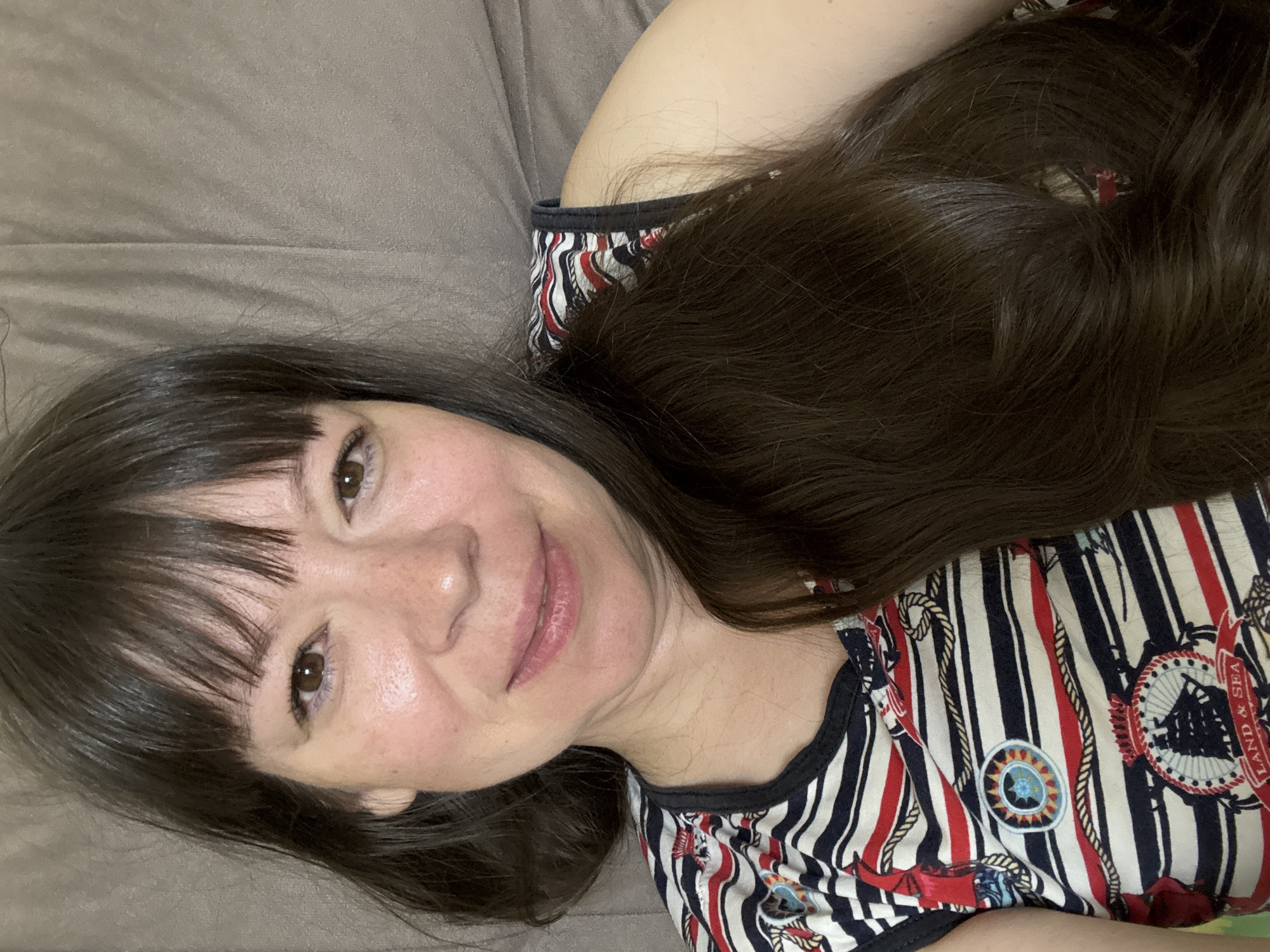 The image shows a close-up, selfie-style portrait of a woman with dark brown hair and bangs. She is wearing a sleeveless top with a nautical-themed pattern featuring stripes, ropes, and ship imagery. The background appears to be a textured, light brown surface, possibly upholstery or a blanket. The woman's face is tilted slightly, and she has a gentle smile. The lighting suggests an indoor setting. The provided crop hints indicate close-ups of her facial features, hair, and clothing. The location of Ветлужский, Russia, is not visually ascertainable from the image itself.
