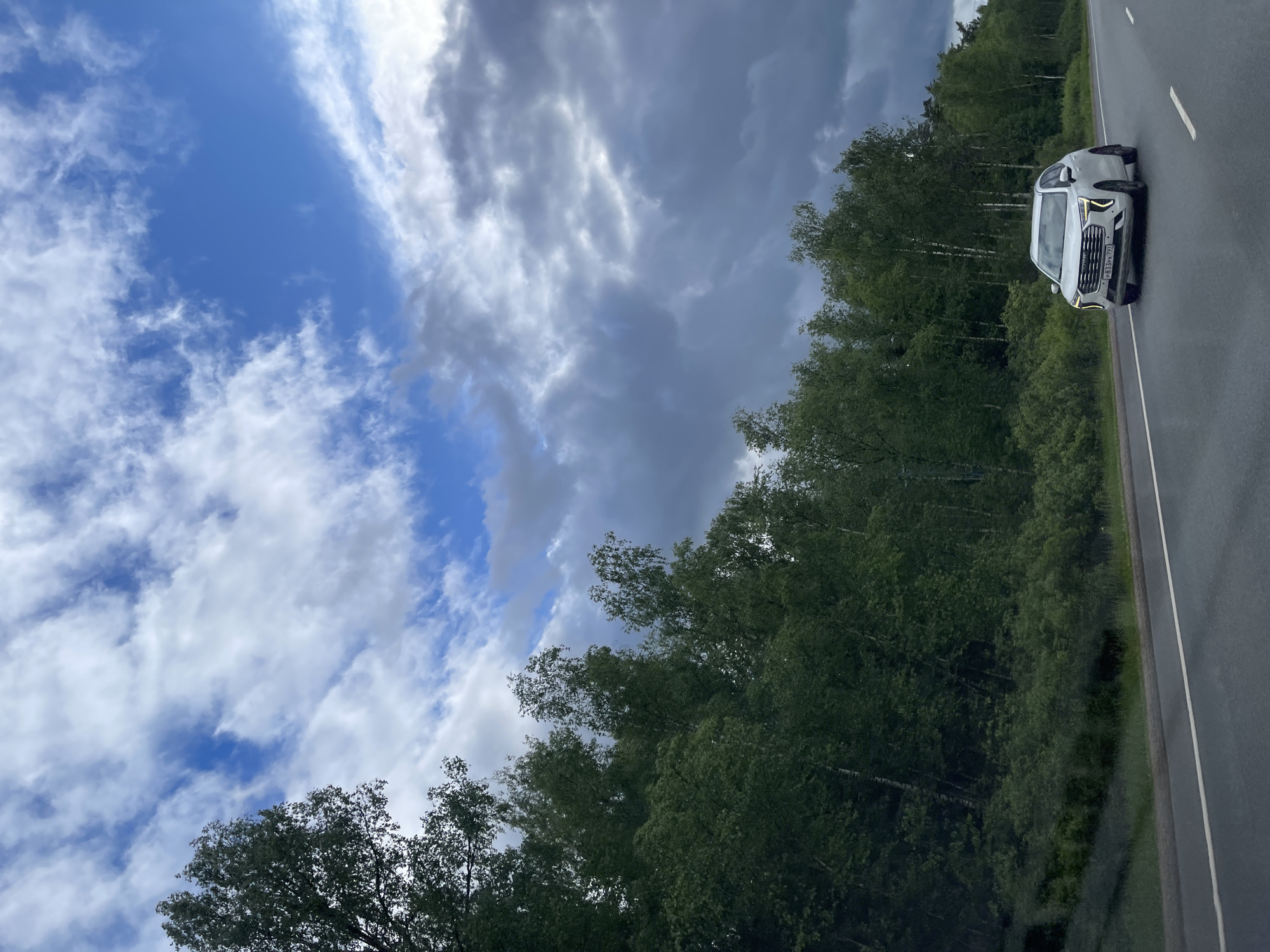 A white car is parked on the shoulder of a paved road in Novyi Byt, Russia. Adjacent to the road is a dense line of trees. The sky above is partly cloudy, with patches of blue visible between the clouds.