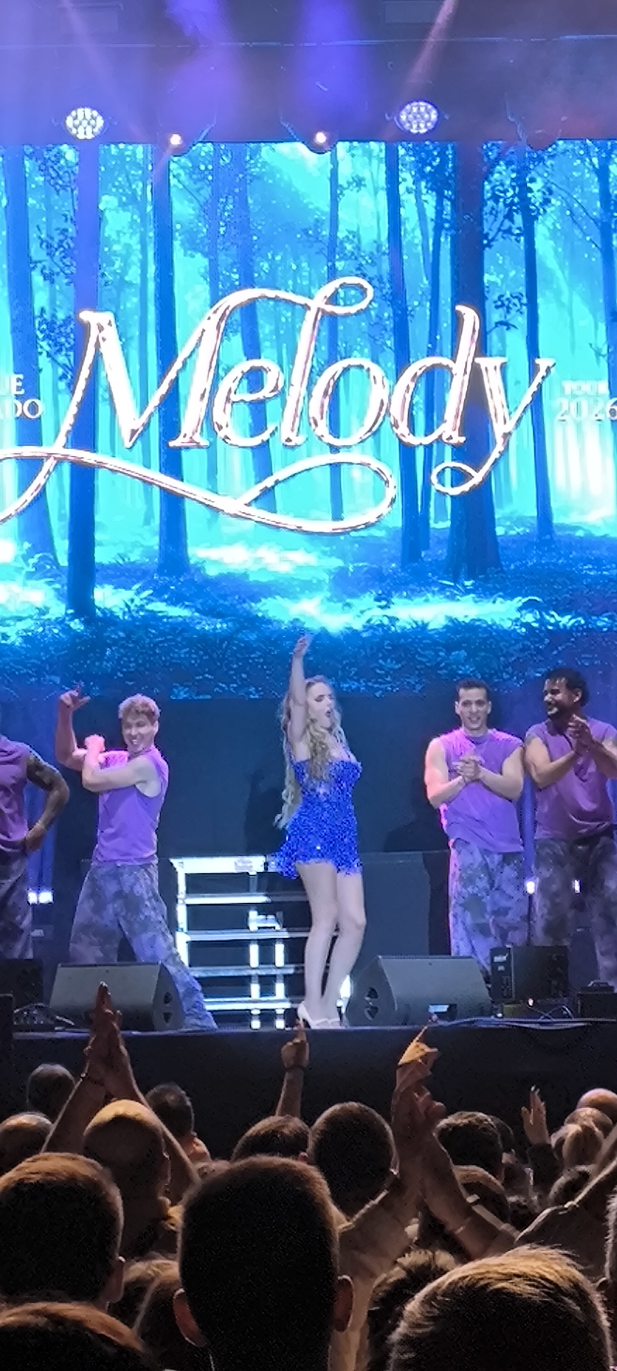 A photograph taken at an outdoor concert in Cerdanyola del Vallès, Spain, depicts a female performer in a blue sequin dress and white high heels on a stage, flanked by four male dancers in purple tank tops and patterned pants. The backdrop features a digitally projected forest scene with the name "Melody" prominently displayed. A crowd of spectators is visible in the foreground, with many raising their hands in applause. Speakers are positioned on the stage floor. The environment suggests an evening or nighttime concert setting with stage lighting illuminating the performers.