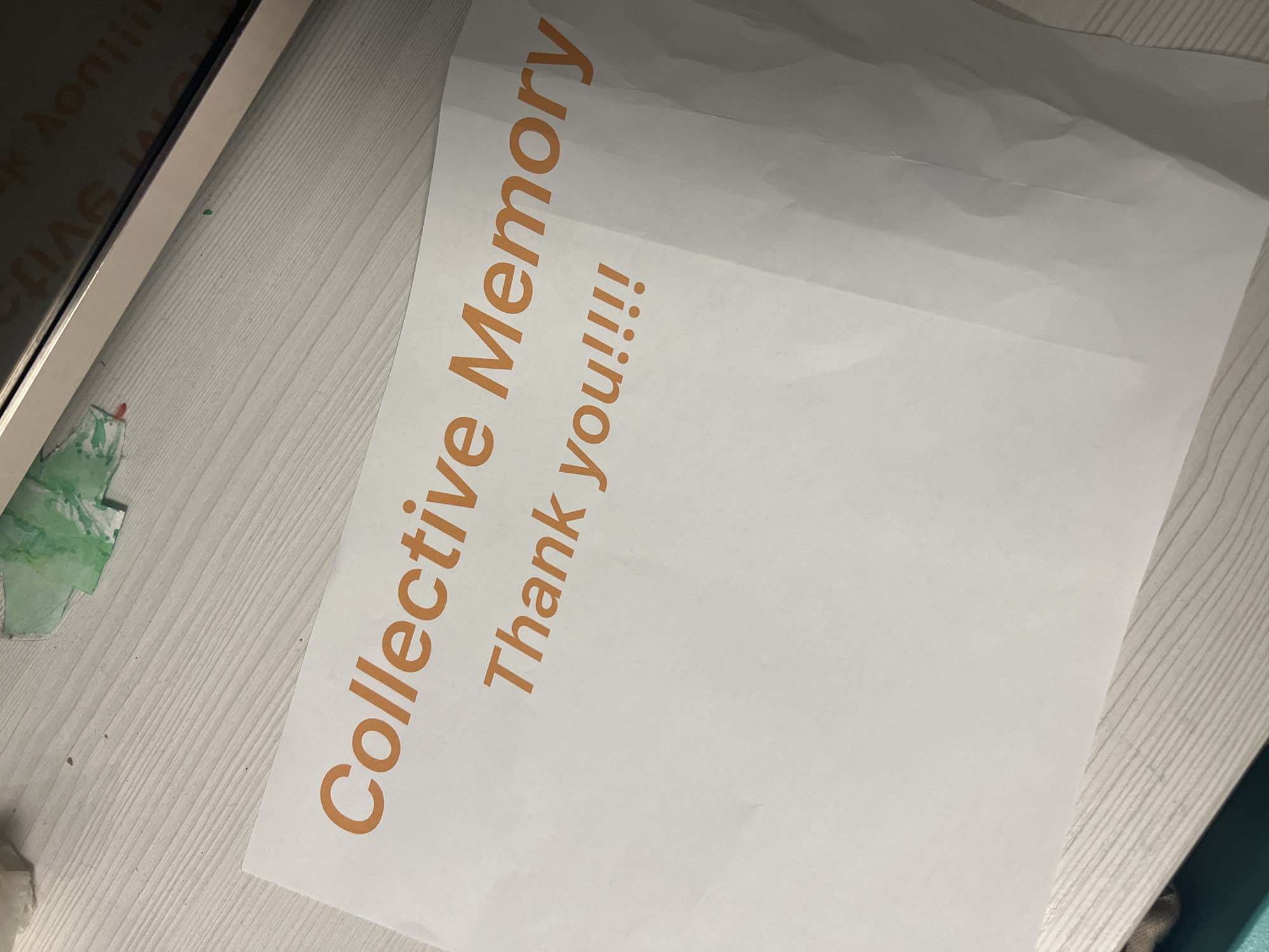 A slightly crumpled white piece of paper is placed on a surface with a light wood-grain pattern. On the paper, printed in an orange-brown color, are the words "Collective Memory" stacked vertically, with "Thank you!!!" below them. To the left of the paper, a portion of a laptop screen is visible, along with a small green, white, and red paper cutout. To the right of the paper is a dark teal surface. The scene is well-lit. Noviy, Russia is not visibly indicated in the image.