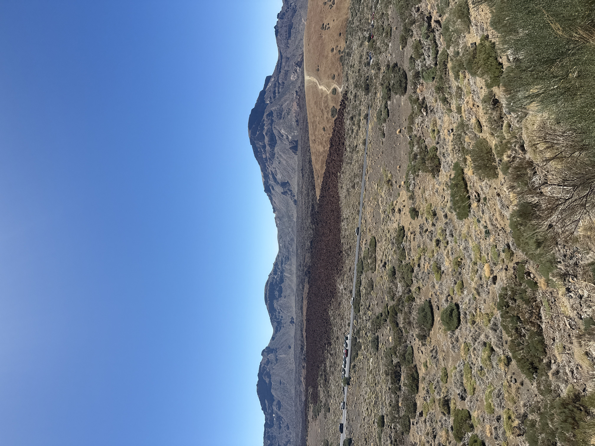 The image shows a vast, arid landscape under a clear blue sky. In the foreground, low-lying scrub and bushes cover the rocky terrain. A road runs horizontally across the middle ground, with several vehicles parked along its side. Beyond the road, a dark, rough expanse of volcanic rock stretches across the landscape. In the background, a range of mountains with rugged, eroded peaks forms the horizon. The overall scene depicts a dry, mountainous environment, likely a volcanic region, under bright sunlight. The location is identified as a viewpoint overlooking the volcanic landscape in La Orotava, Spain.