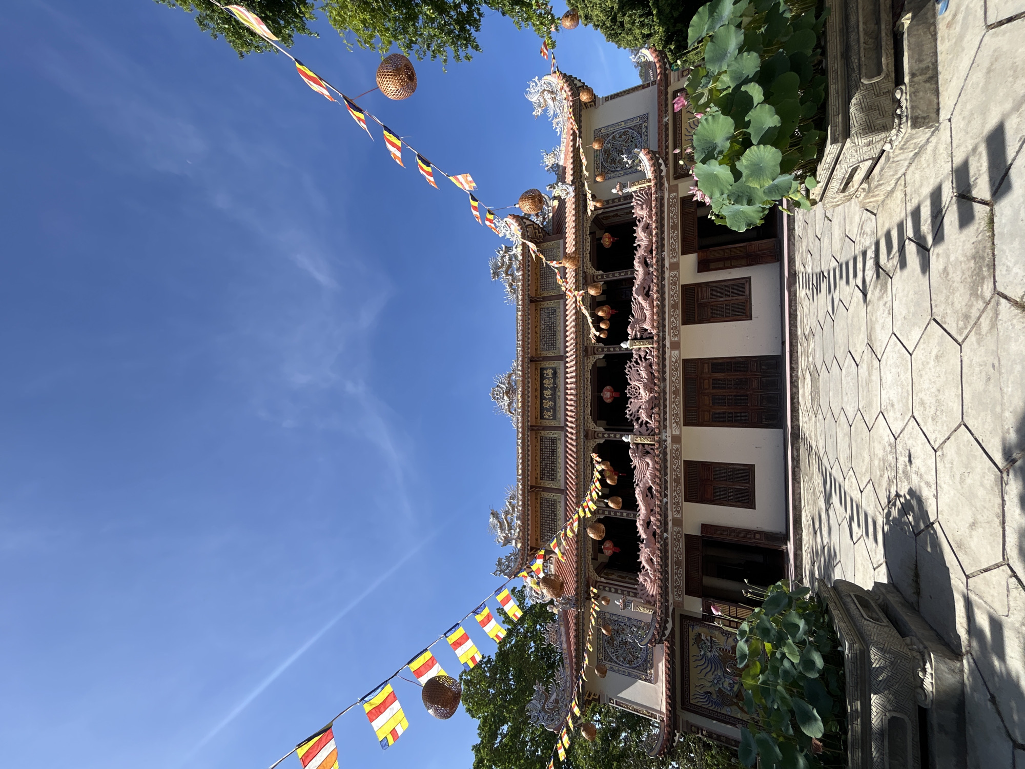 The image shows a temple in Tay Nha Trang Ward, Vietnam, under a clear blue sky. Colorful Buddhist flags and decorative woven lanterns are strung across the building's facade. The temple has multiple tiers with intricate dragon carvings and architectural details. The ground is paved with hexagonal stones. A cluster of lotus plants grows near the entrance.