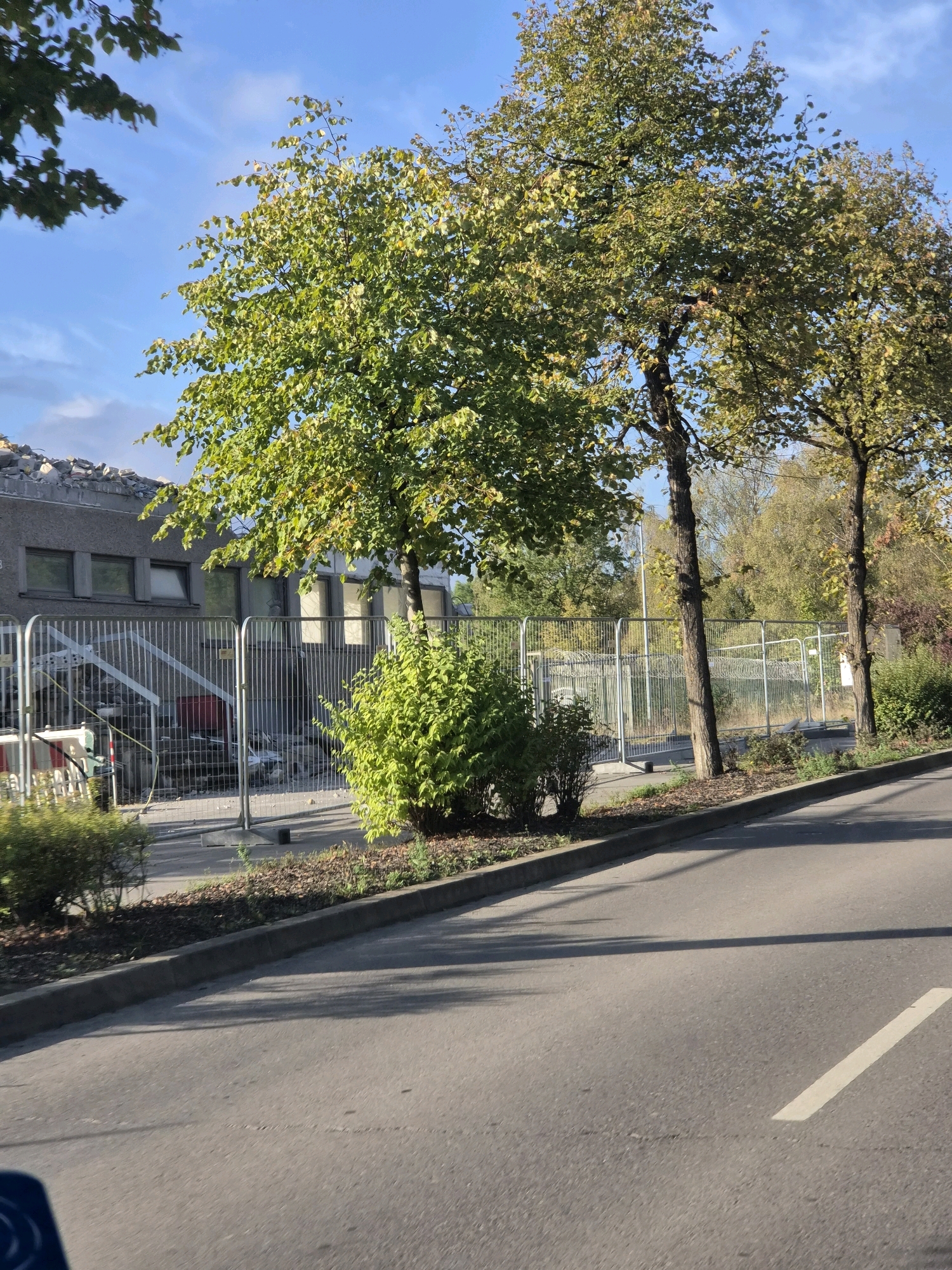 The image depicts a street scene in Leudelange, Luxembourg, on a clear day. In the background, a partially demolished building is visible, with debris on its roof. A metal fence surrounds the construction site, separating it from the street. Trees with green and yellowing leaves line the street, casting shadows on the asphalt. The road in the foreground appears to be an active thoroughfare, with a white line marking a lane. No individuals are present in the image.