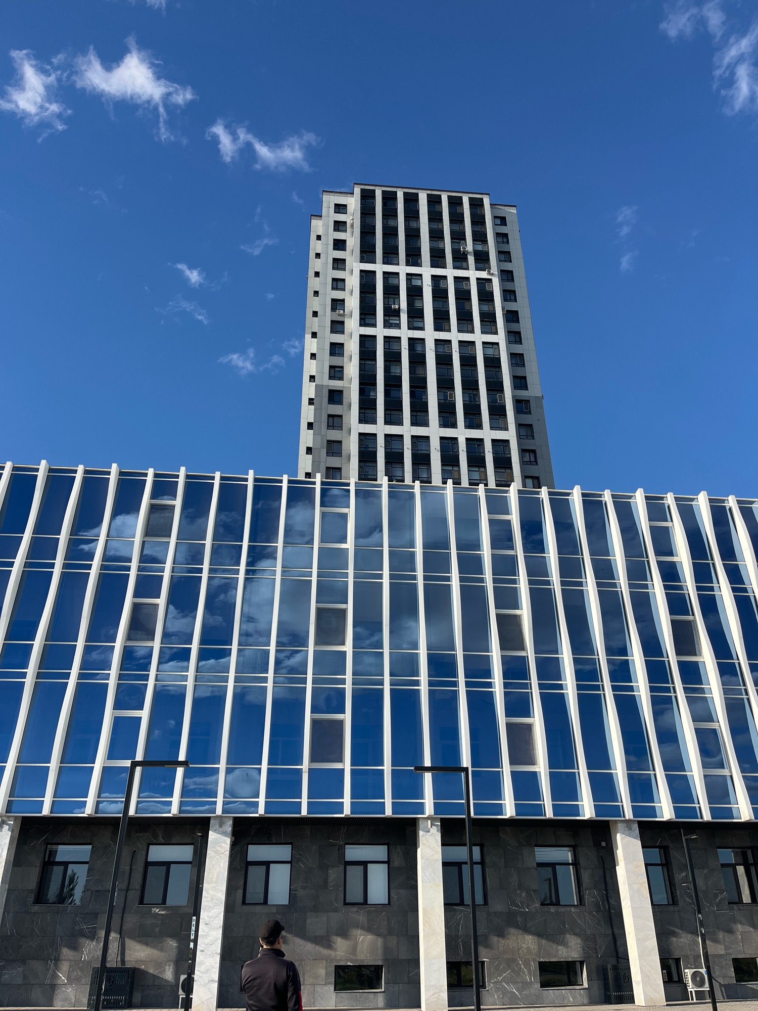 The image shows a modern building with a glass facade and a tall residential tower against a bright blue sky with scattered clouds. The building's glass reflects the sky and clouds, creating a dynamic visual effect. The lower part of the building features dark, textured stone panels and several dark-framed windows. A solitary individual wearing a black jacket and a black cap stands with their back to the camera, facing the building. A black lamppost stands in front of the building. The scene appears to be in a contemporary urban setting, potentially in Astana, Kazakhstan, given the architectural style.