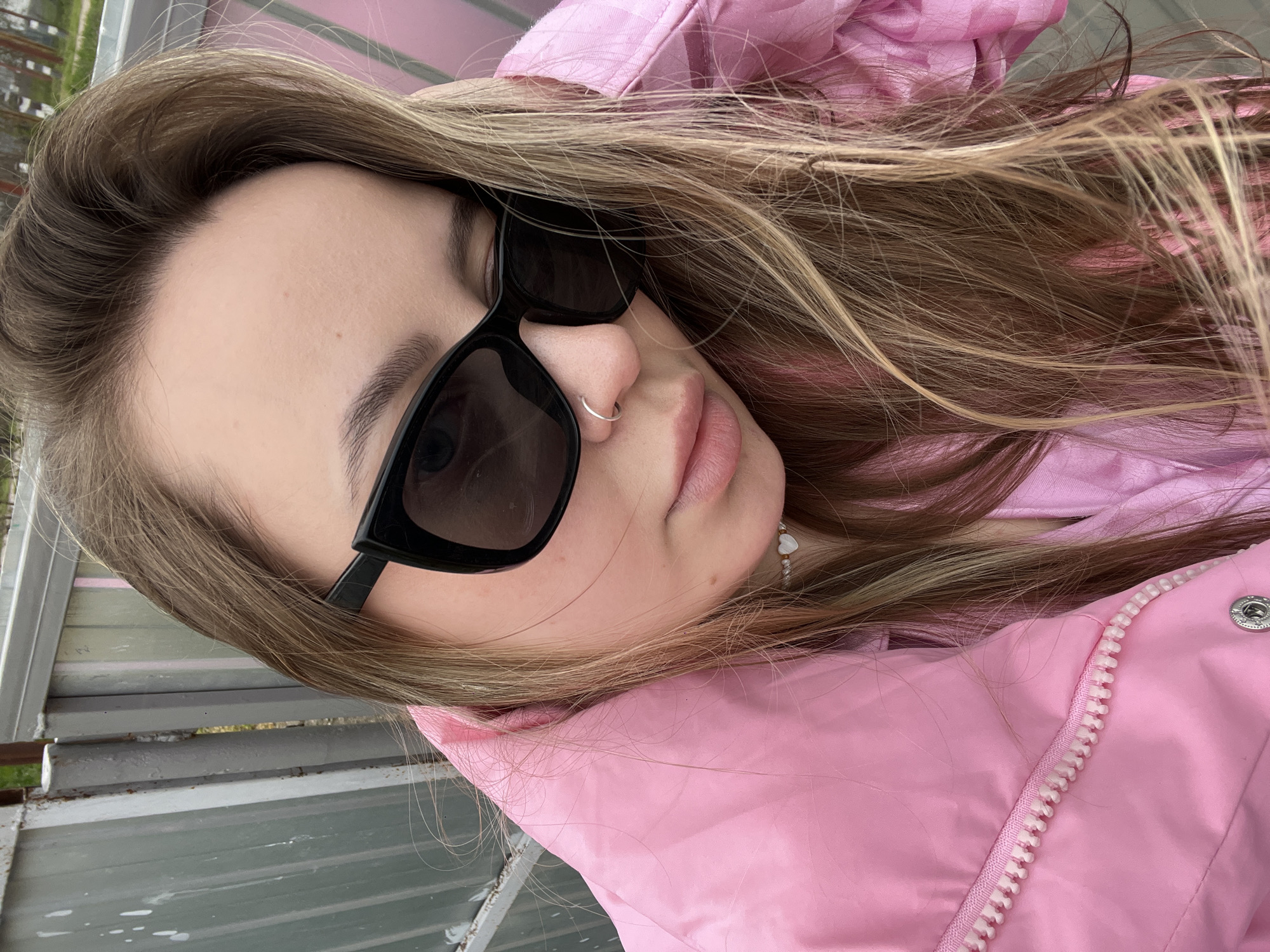 A young woman with long, brown hair and sunglasses is wearing a pink jacket and standing outside in what appears to be Novopokrovskaya, Russia. She has a nose ring and a necklace with a heart charm. The background shows a grey metallic structure, potentially a fence or building component.