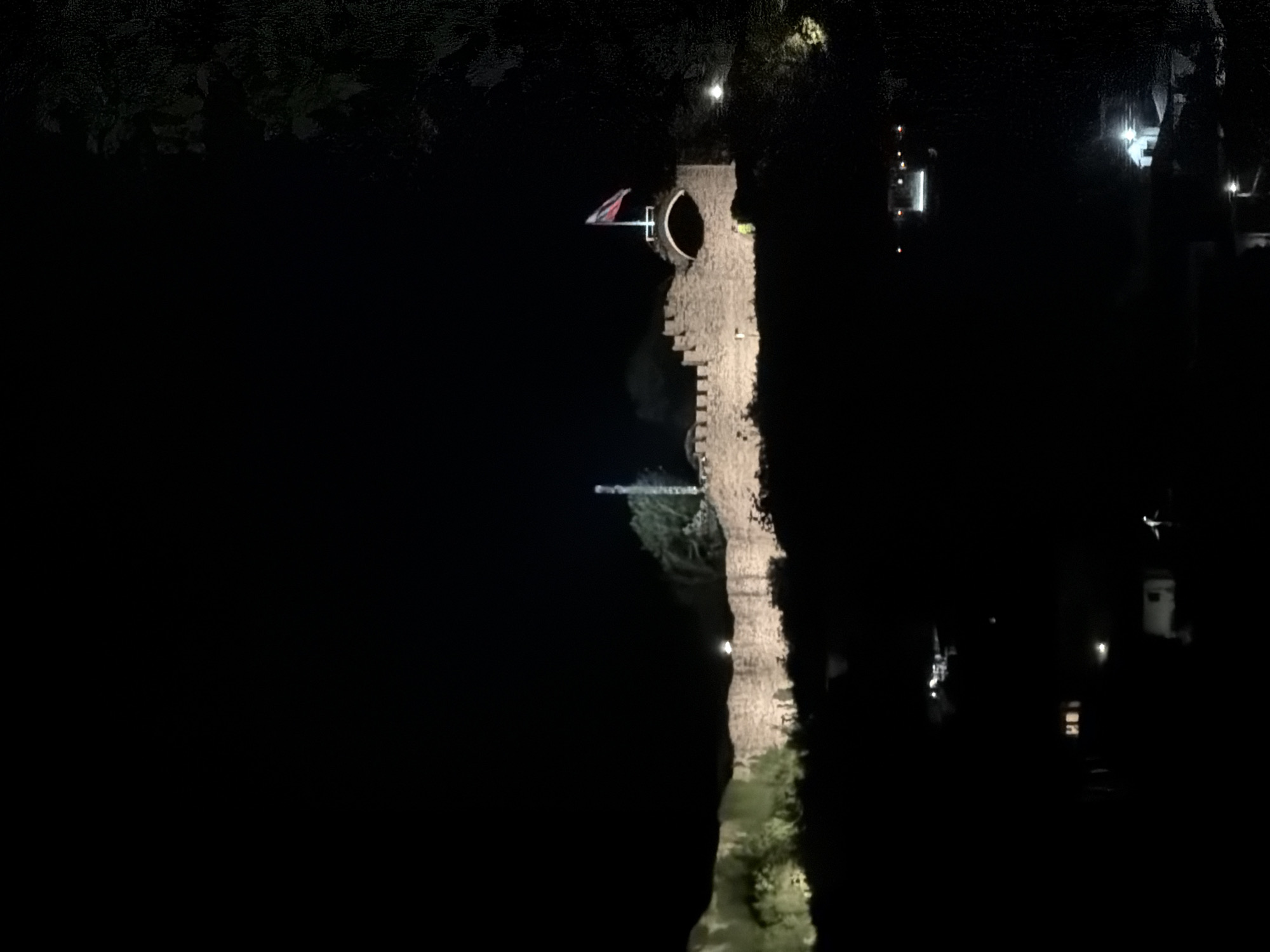 An aerial, night view depicts a castle illuminated in the darkness of Unknown, Unknown. The castle tower stands tall with a flag at the top, appearing medieval. The scene is almost completely dark, with light sources focusing on the castle itself and a few surrounding structures. The landscape around the castle is undefined due to the darkness.