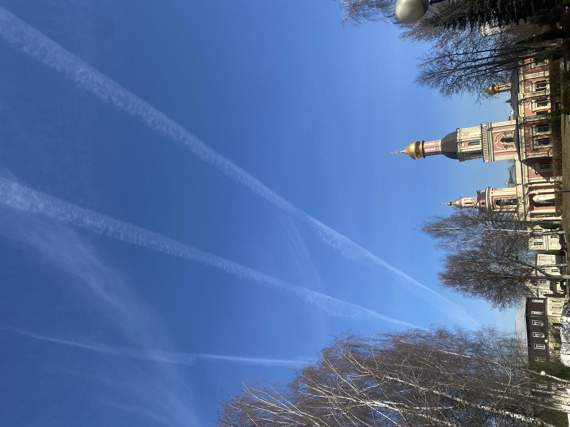 The image depicts a clear blue sky with multiple contrails crisscrossing above a building, identified as the Novodevichy Convent in Moscow, Russia. The convent features a golden dome and a multi-story structure with red and white facades. Bare trees are visible in the foreground, framing the view of the building and sky. There are also what appear to be pink or reddish objects among the contrails.