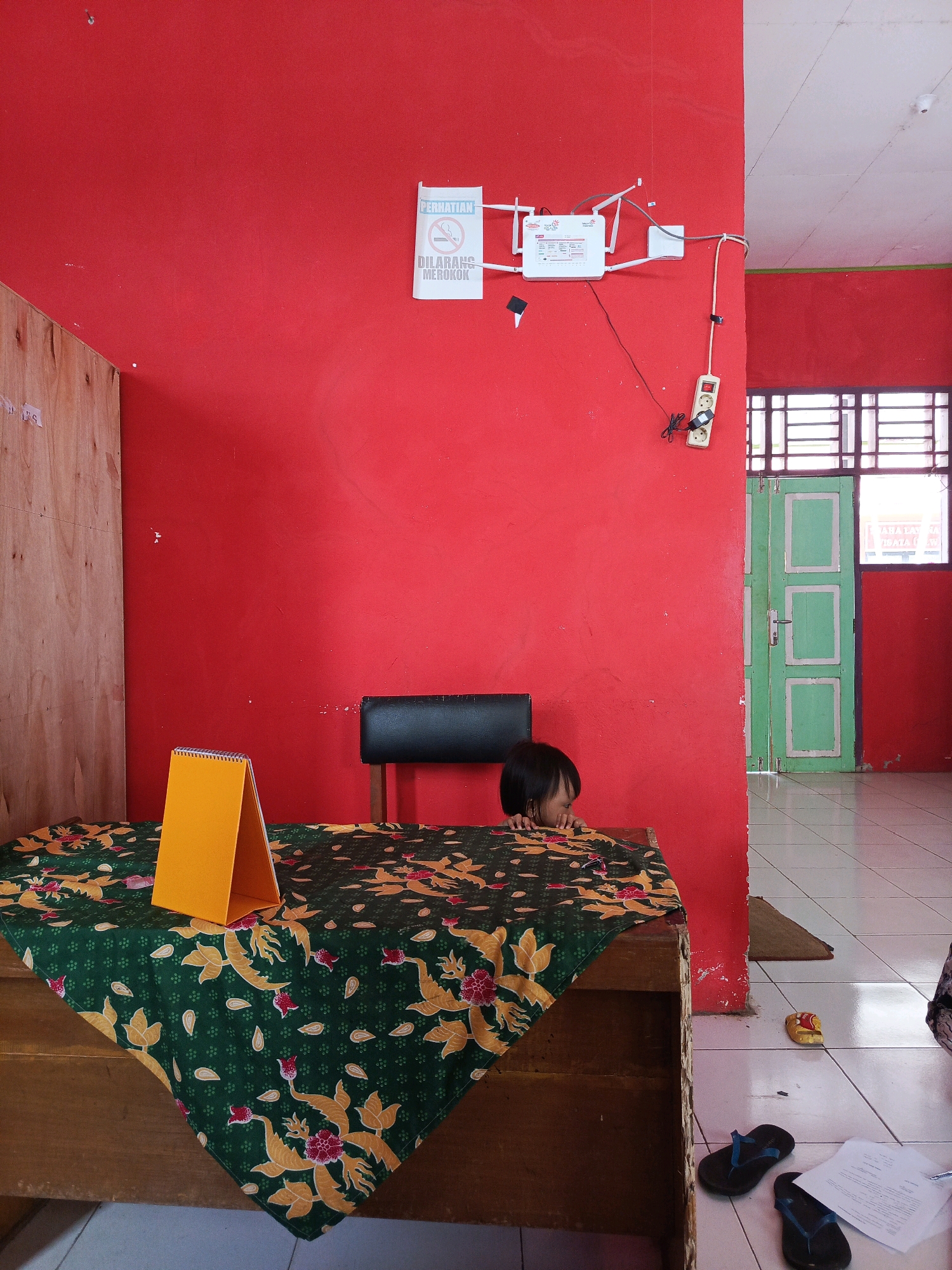 The image depicts an indoor scene with bright red walls. In the foreground, a wooden table is covered with a dark green tablecloth featuring a floral pattern in yellow and red. An orange, tent-shaped object, possibly a desk organizer or a small stand, is placed on the tablecloth. To the right of the table, a child with dark hair is partially visible, peeking over the edge of the table. Behind the child, a black chair is present. Further to the right, a green door with white trim is visible, leading to an area with tiled flooring. On the red wall, a white sign with the text "PERHATIAN DILARANG MEROKOK" (Attention No Smoking) is affixed. A white Wi-Fi router with multiple antennas is also mounted on the red wall, connected by wires to an electrical outlet. A power strip with two sockets is plugged into the wall. The overall environment suggests a modest, possibly public or administrative space within a building in Tik Jeniak, Indonesia.