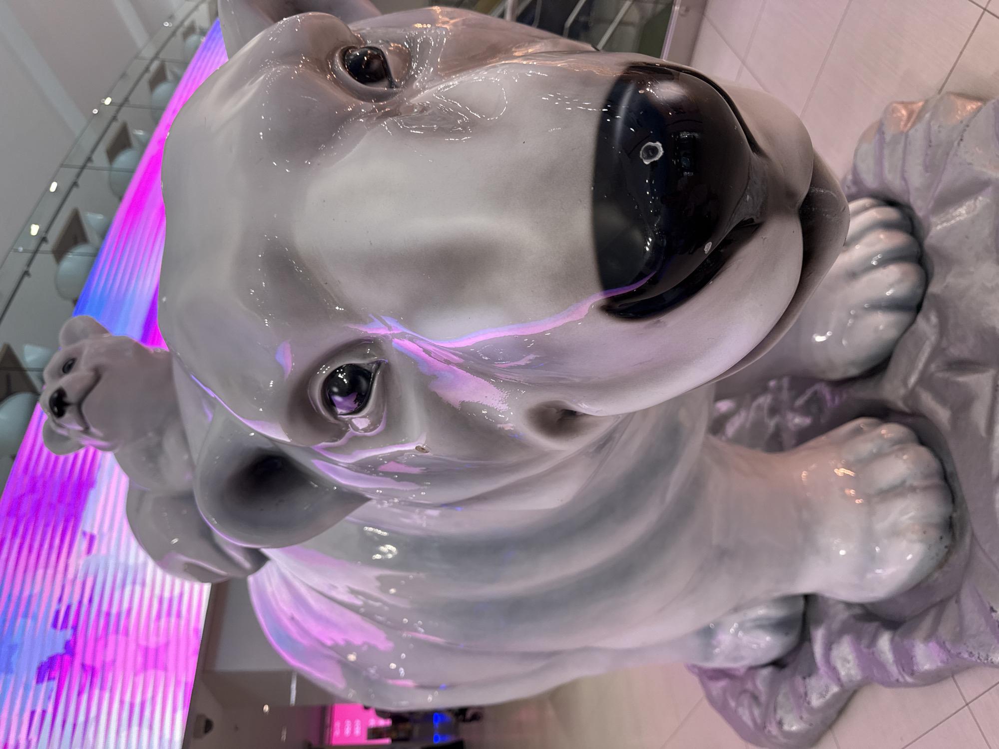 The image shows a glossy, white, ceramic polar bear sculpture, possibly located in a public space, likely a shopping mall. The bear is depicted in a seated position, with its front paws visible. A smaller bear cub sculpture is visible, seemingly nestled against the mother bear. The lighting in the area appears to be fluorescent, with a purple and pink hue reflected on the bear's surface from a nearby light source, possibly a LED display. In the background, there's a glimpse of the mall's architecture, which includes metallic structures and lighting fixtures. The sculpture's surface has some visible imperfections, like streaks and spots, suggesting it's been touched or cleaned. There is no sign of people. The location within Moscow, Russia, cannot be determined with certainty from the image.