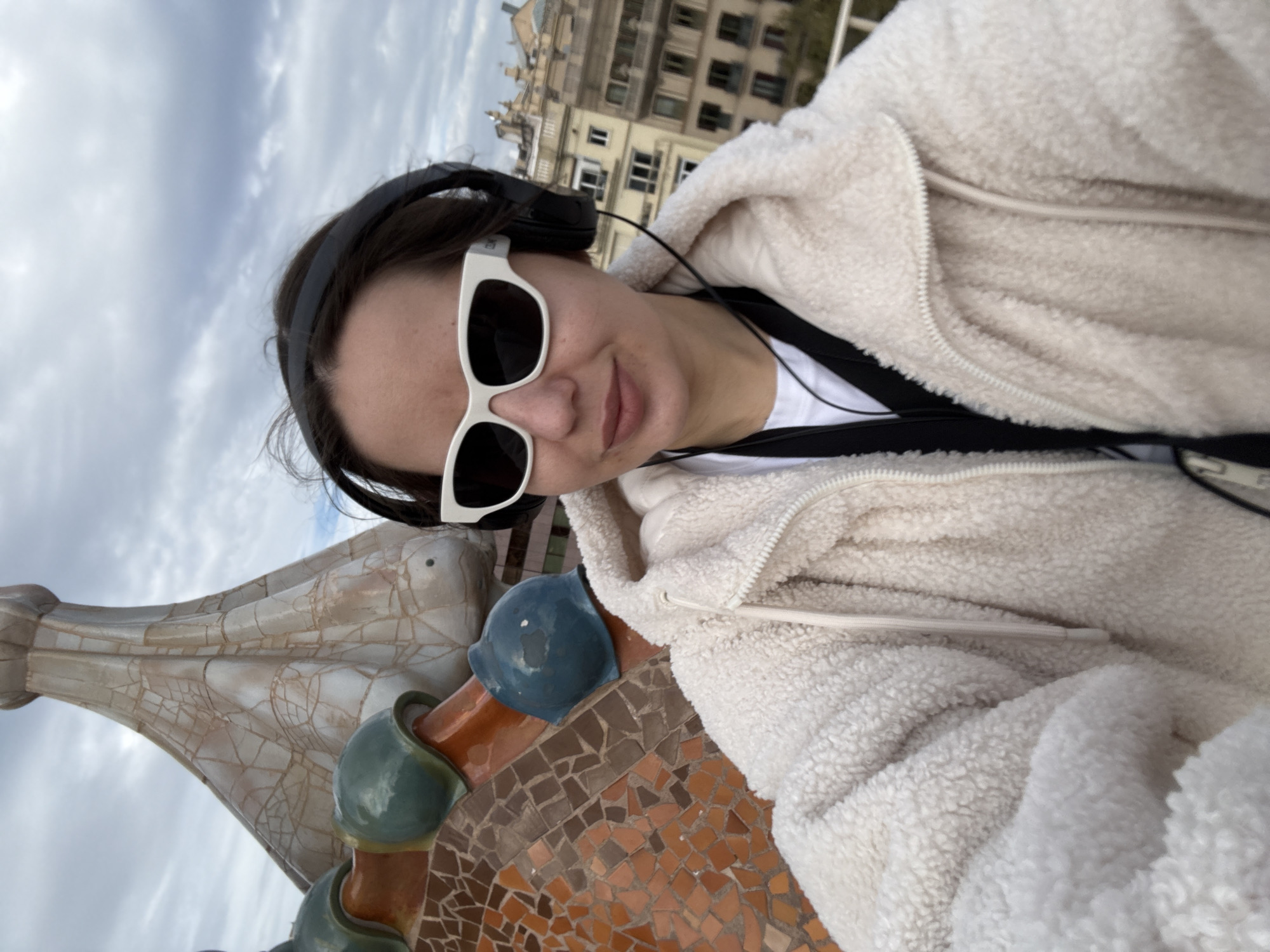 A female subject is centered in a close-up selfie, positioned on the rooftop of Casa Batlló in Barcelona, Spain. She has dark hair, wears white-framed sunglasses with dark lenses, and black over-ear headphones with a visible cord. Her attire includes a white t-shirt and a cream-colored, textured fleece jacket with a visible zipper and drawstring. A black strap is visible across her chest. Her expression is neutral.
Behind her, architectural elements characteristic of Casa Batlló are prominent. To the left and behind her head, a large, organic-shaped chimney rises, displaying a light beige color with a distinct cracked pattern texture. Below the chimney and to the subject's left, a section of the roof's "dragon's back" structure is visible, composed of colorful mosaic tiles (trencadís) in shades of orange and brown, interspersed with rounded, teal and blue scale-like decorative elements. In the distant background, typical multi-story urban buildings of Barcelona are visible under an overcast sky.