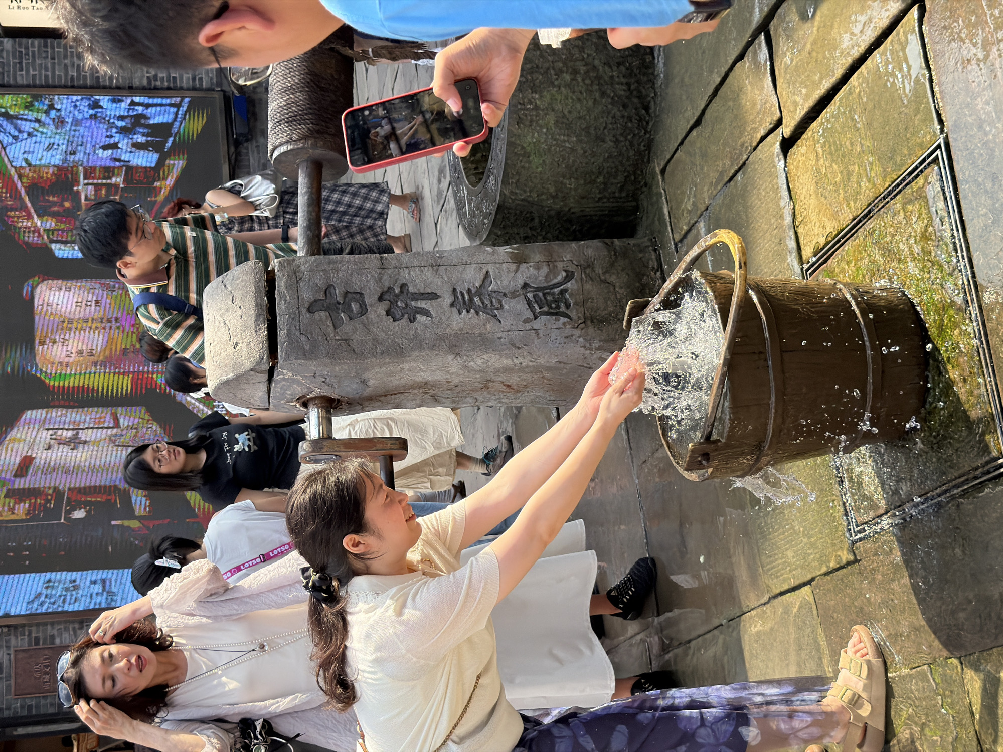 In the Yuzhong District of China, a woman in a cream-colored top and patterned skirt bends down, splashing water from a wooden bucket. Her hands are wet, and water is spraying from the bucket. Behind her, other people are visible. To the left, a woman wearing sunglasses and a light cardigan stands, touching her hair. Further back, a woman in a black t-shirt and glasses looks to the side, and a man in a striped shirt and glasses leans forward, looking towards the well structure. A red smartphone is held up by a person on the right, seemingly taking a picture of the scene. The environment is paved with stone slabs, and there's a well with carved characters on its stone structure. The scene appears to be outdoors in a public area with a large digital display in the background.