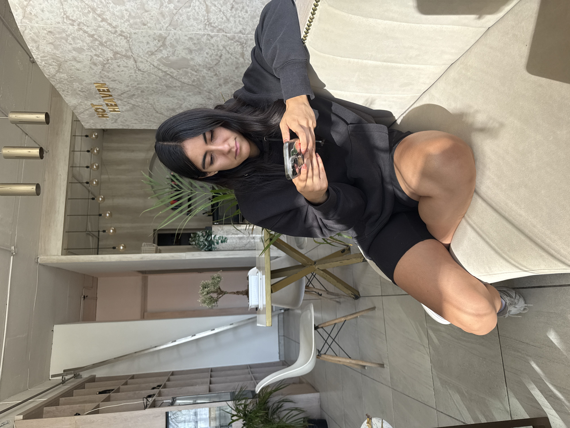 A young woman with long, dark hair sits on a light-colored sofa in a modern indoor setting, likely in Puebla, Mexico. She is dressed in a black hoodie and black athletic shorts, with light-colored sneakers visible on her feet. Her gaze is directed downwards at a smartphone she holds in both hands, appearing focused and calm as she interacts with the device, which has a clear case showing a floral pattern.
The interior space features light-colored tiled flooring and walls. A prominent wall behind her has a marble-like texture, adorned with the golden, raised lettering "HOT HEAVEN." Overhead, modern metal cylindrical light fixtures hang from the ceiling, and several bare bulb lights are visible in the background. Bright natural light streams in, casting sharp shadows from furniture onto the floor, indicating daytime. The background also includes a small table with a white chair, shelving units, and various potted plants, contributing to a contemporary and airy atmosphere.
