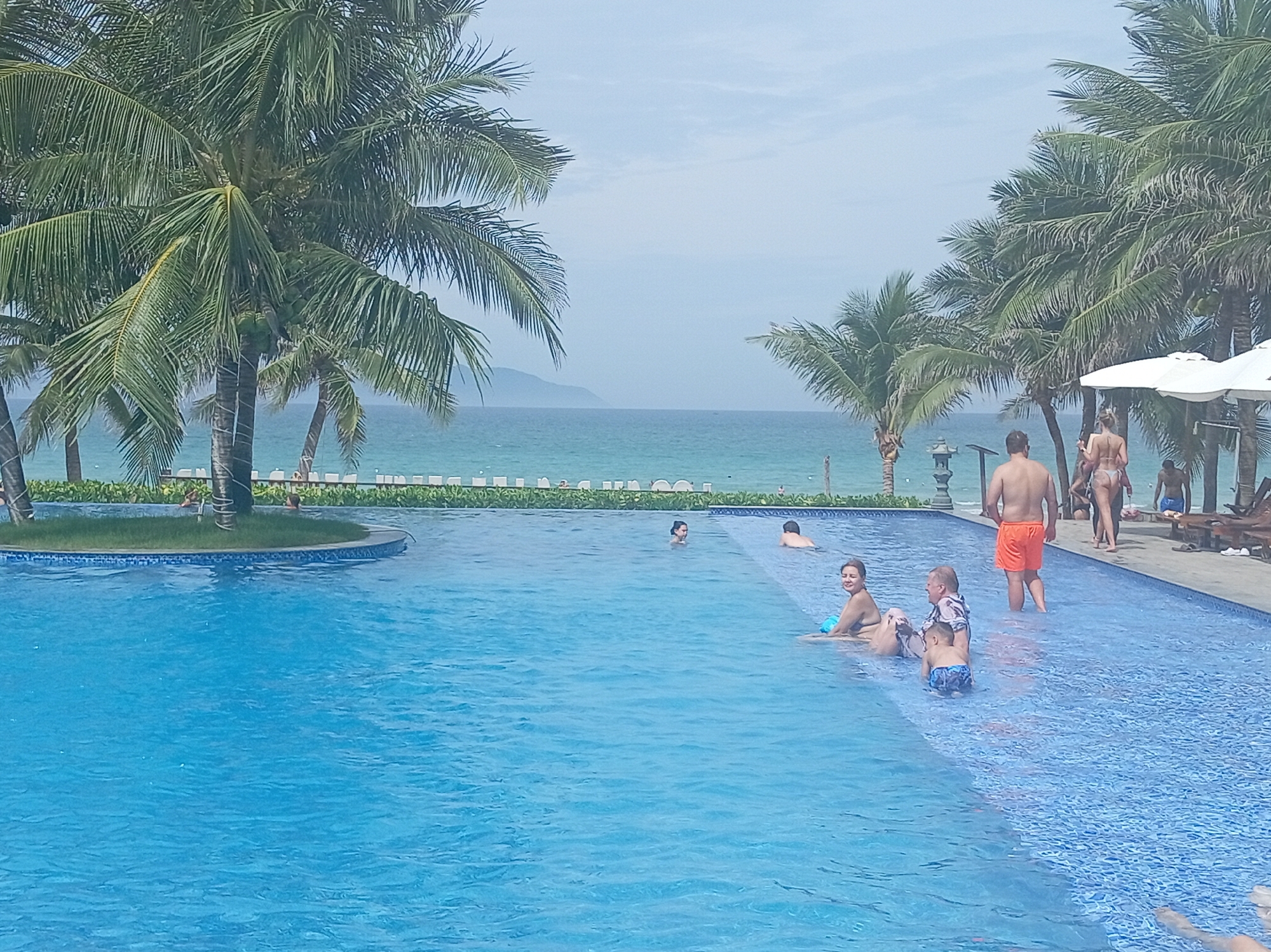 In Dong Ninh Hoa, Vietnam, a large, crystal-clear blue swimming pool is the central focus of this scene. The pool is adjacent to a tropical beach with lush palm trees framing the view. The ocean, a vibrant turquoise, stretches out to the horizon, meeting a hazy, pale blue sky. Several people are enjoying the pool and its surroundings.
In the foreground, the water is a deep, inviting blue, with intricate mosaic tiles visible on the pool floor. Several individuals are in the pool: a woman with dark hair is sitting on the edge, leaning back and looking to her left; a man in orange shorts stands in the shallow end, facing away from the camera; and a young boy in blue swim trunks is playing in the water near the edge. Further in the pool, a couple, a woman in a bikini and a man, are interacting.
On the far side of the pool, a paved area is lined with lounge chairs and large white umbrellas, suggesting a resort or hotel setting. Several people are visible on this paved area, including a woman in a bikini and other figures in the distance. The background reveals a verdant landscape with more palm trees and the distant mountains of Vietnam.