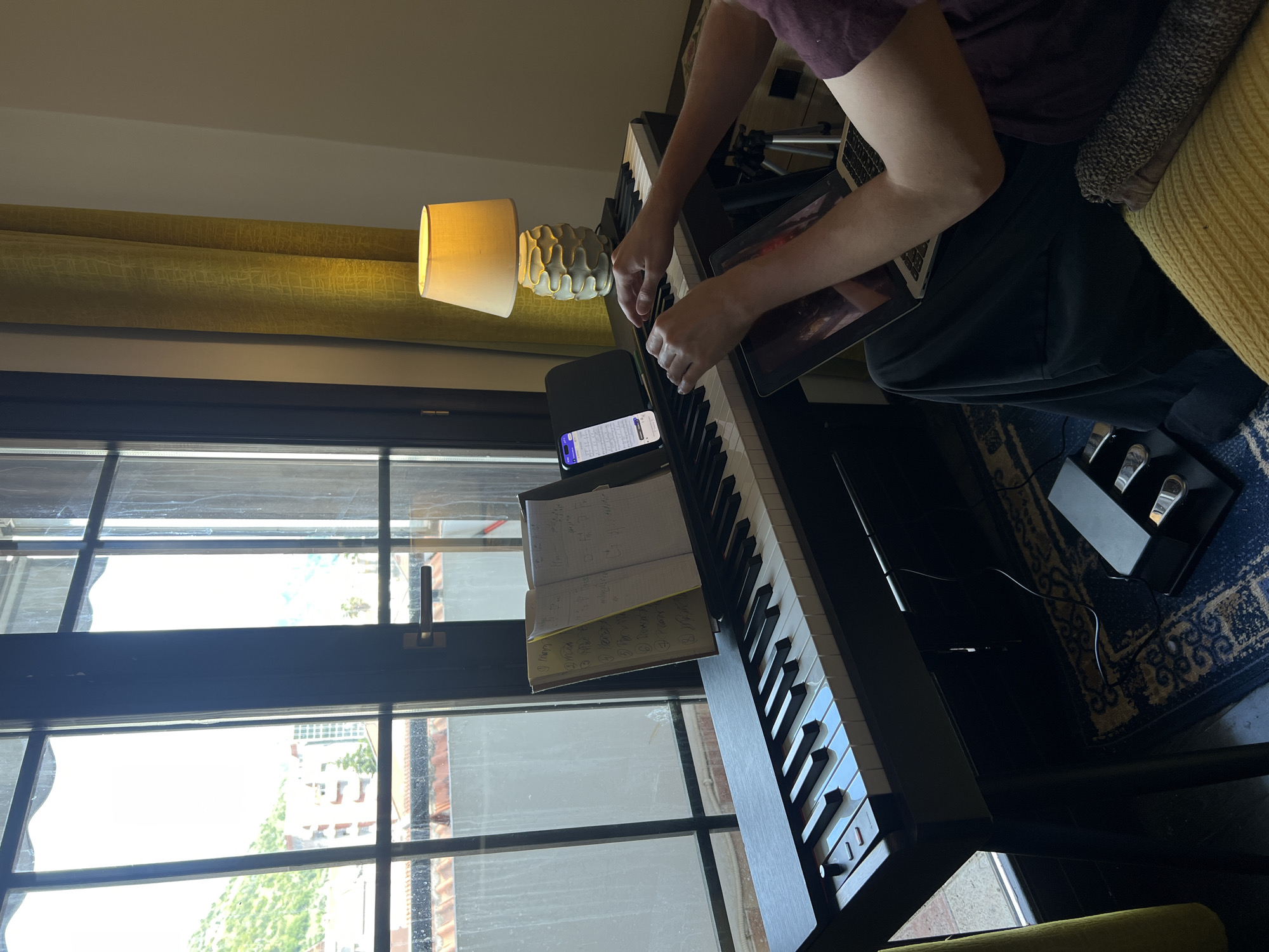 The image depicts an indoor scene in Budva, Montenegro, featuring a person engaged in playing a digital piano during what appears to be daytime. The person, seen from the torso upwards, wears a dark red t-shirt and dark pants, with their hands positioned over the black and white keys of the piano.
The digital piano is central to the image, with three chrome pedals visible on a dark blue patterned rug below it. An open notebook, serving as sheet music, rests on the piano's stand, displaying handwritten musical notations, including chord symbols like "Am," "Dm," "G," and "C/F," alongside a numbered list that appears to include song titles or exercises such as "Norah," "Wicked," "Blue," "Rain," "River," and "Dreams." Next to the notebook, a smartphone on a stand shows a screen with text, possibly from a messaging or list application. On the person's lap, a laptop or tablet is open, displaying an image on its screen.
To the left of the piano, a table lamp with a cream-colored shade and a textured ceramic base emits a warm glow. Behind the lamp, yellow textured curtains are drawn. A large window dominates the left side of the frame, letting in bright natural light and offering a view of an outdoor urban environment with buildings and some greenery under sunny skies. The setting suggests a dedicated space for musical practice or leisure.