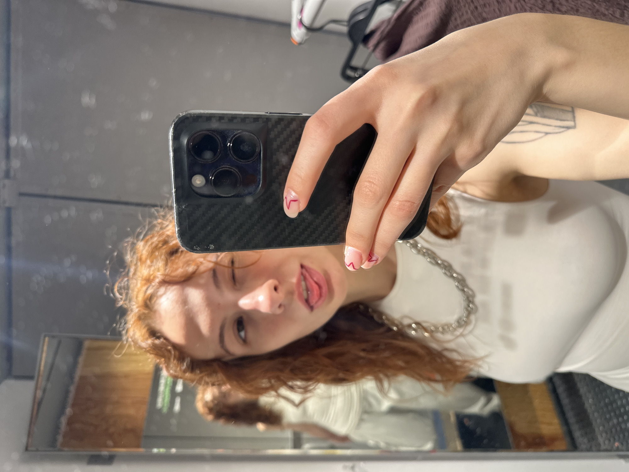 A young woman is taking a selfie in a mirror. She has red hair, fair skin, and is wearing a white tank top and a silver chain necklace. Her nails are painted with a minimalist red design. She is holding a phone with a carbon fiber case, obscuring part of her face. Her reflection is visible in the mirror. The setting appears to be an indoor location, possibly a dressing room, with clothes visible in the background. The specific location in Novi Sad, Serbia is not discernible from the image.