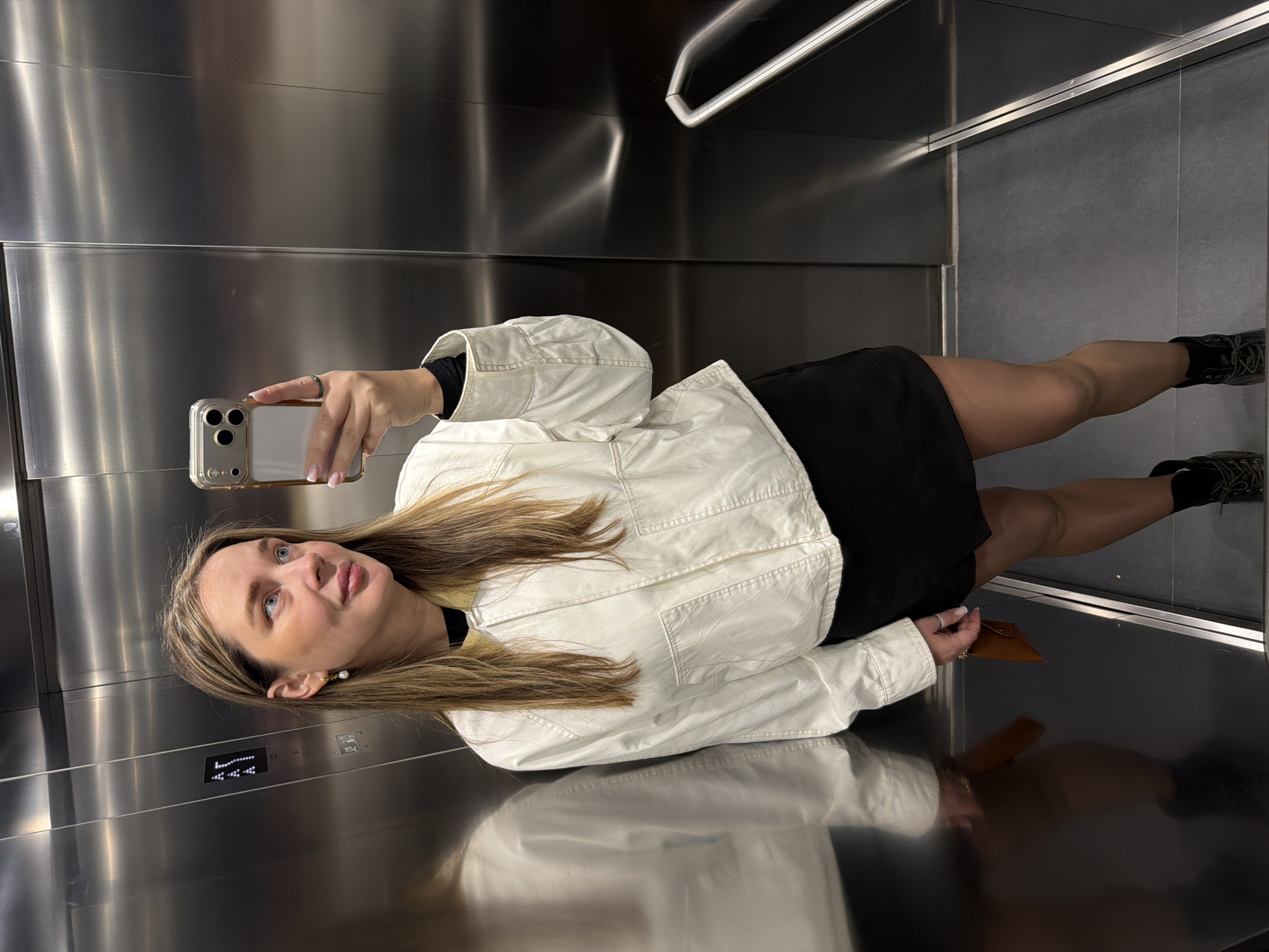 A woman is taking a selfie in an elevator. The elevator has stainless steel walls and a grey floor. The woman is wearing a black dress, a white jacket, and black boots. She is holding a cell phone in her right hand. The elevator display shows the number 14.