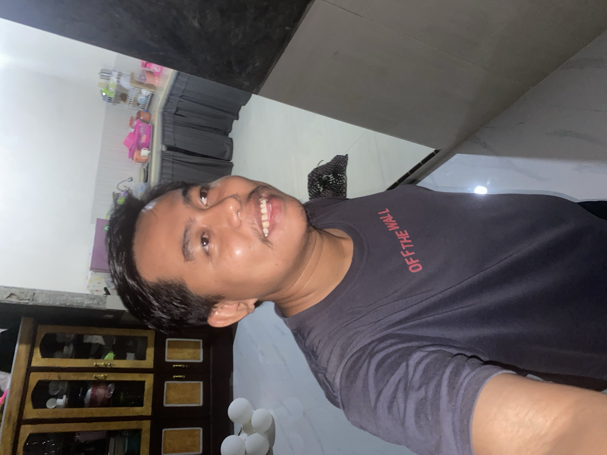 A man in a dark blue t-shirt with the red text "OFF THE WALL" is taking a selfie in what appears to be an indoor kitchen or dining area. He has dark hair, a mustache, and is smiling with his teeth visible. In the background, there is a wooden cabinet with glass doors, kitchen cabinets with a dark curtain, a sink, and various kitchen utensils. There are also white balloons visible to the left of the man. The floor is tiled with a marble pattern. The overall scene suggests a casual, domestic setting.
