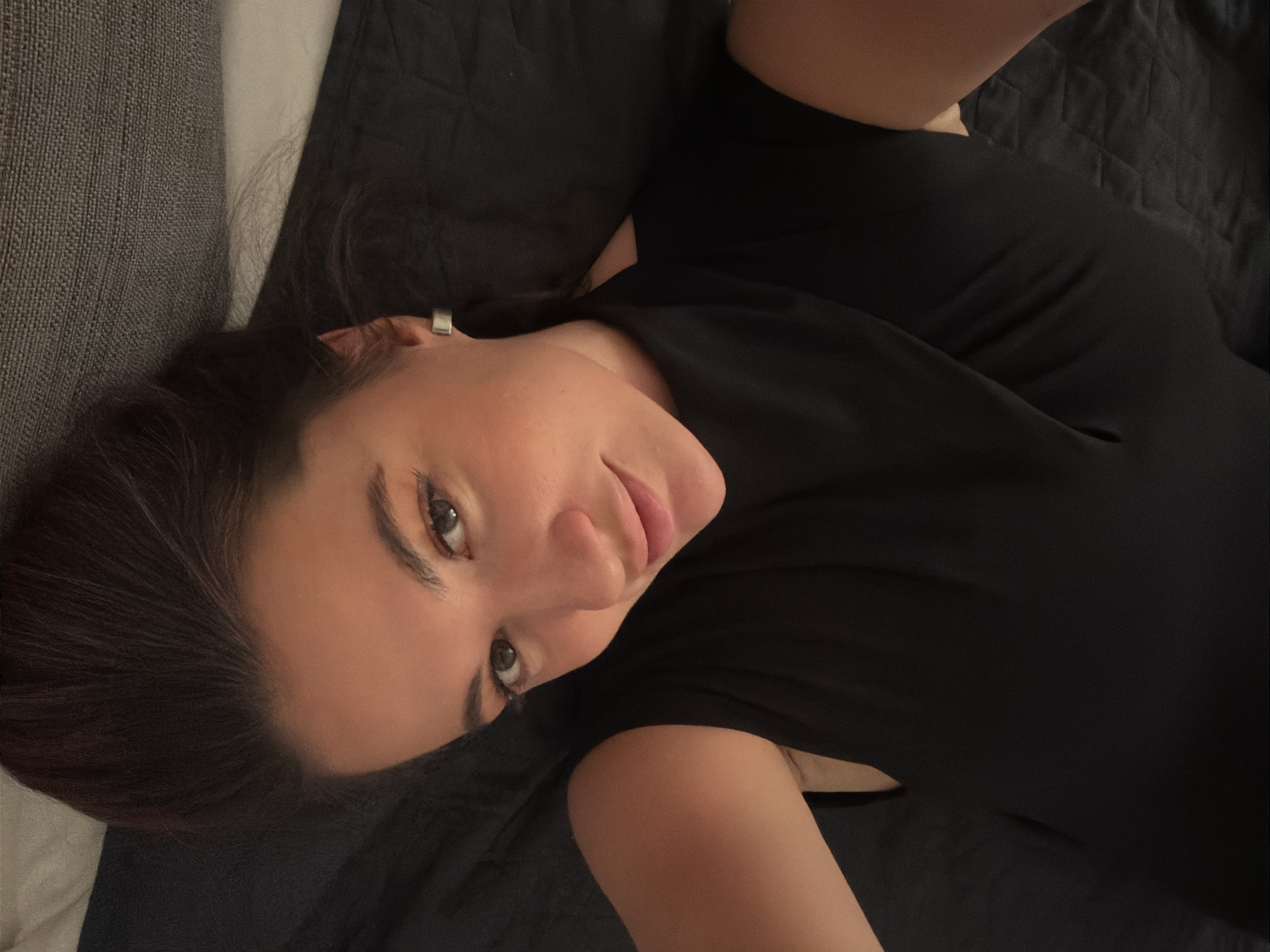 A female individual is depicted in a close-up, upward-facing shot, lying on her back. She wears a form-fitting, short-sleeved black top or dress. Her head is positioned on a dark, quilted fabric surface, with darker hair visible spread out behind her. To the lower left, a lighter grey, textured fabric surface is visible. The individual has a direct gaze towards the camera, with a slight tilt to her head. Her facial features include full lips and dark eyes. No architectural elements, landmarks, or other specific environmental cues are present to indicate a location within the city of Belgrade, Serbia, or any other specific geographic location. The lighting is low and even.