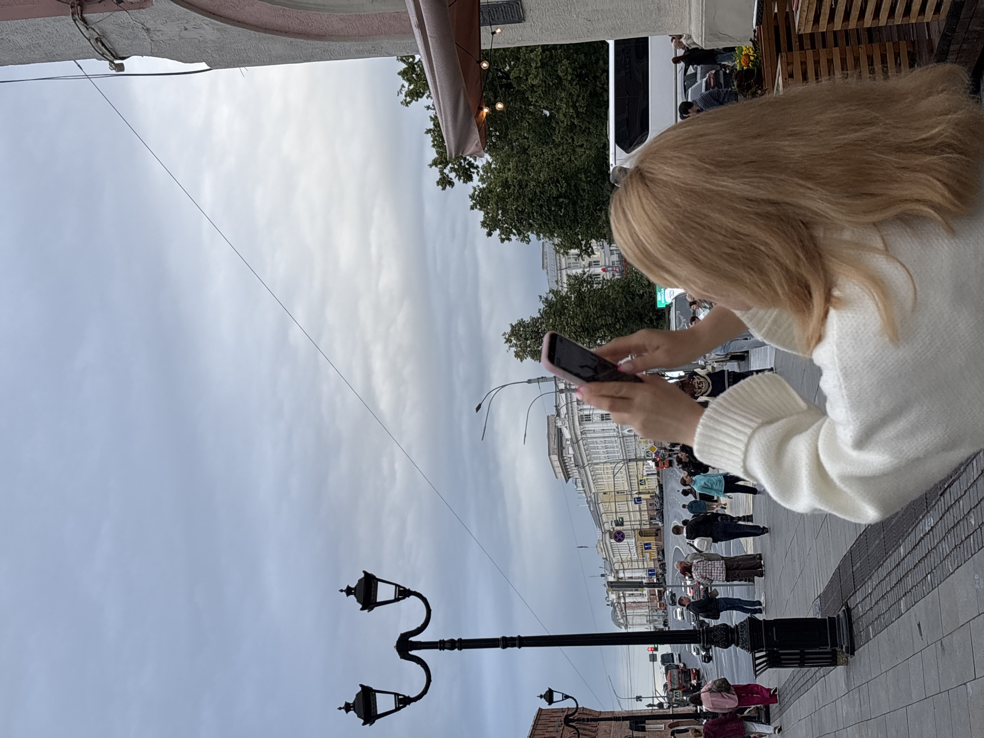 This is a vertical, medium-angle, outdoor shot of a woman with blonde hair wearing a white knitted sweater, holding a pink smartphone and seemingly taking a picture. Her face is not visible. The scene is a street in Nizhny Novgorod, Russia. In the background, there are people walking on the sidewalk, a street, and buildings. A black double-headed lamppost is prominent in the foreground. The sky is overcast, suggesting a cloudy day. The overall impression is that of a candid moment of a tourist or resident capturing a scene in the city. There is no visible text.