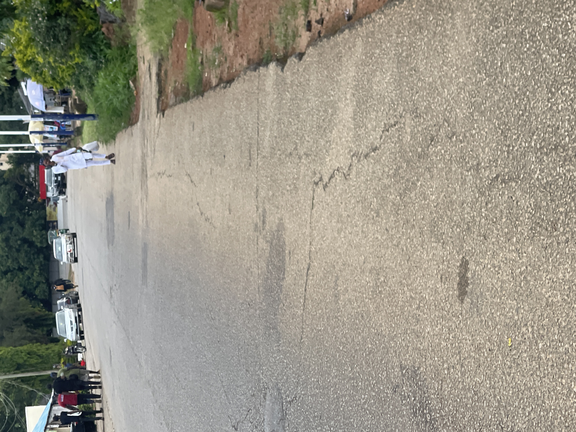 The image shows a street scene in Jos, Nigeria. In the foreground, a grey asphalt road with visible cracks occupies most of the frame, with a concrete sidewalk on the right. A person in white traditional attire is walking on the sidewalk carrying a white bag. Further up the road on the left, several people are standing near a row of parked cars. Among them, four individuals are walking away from the camera. The environment appears to be a street with buildings and vegetation lining the background. The lighting suggests it is daytime.