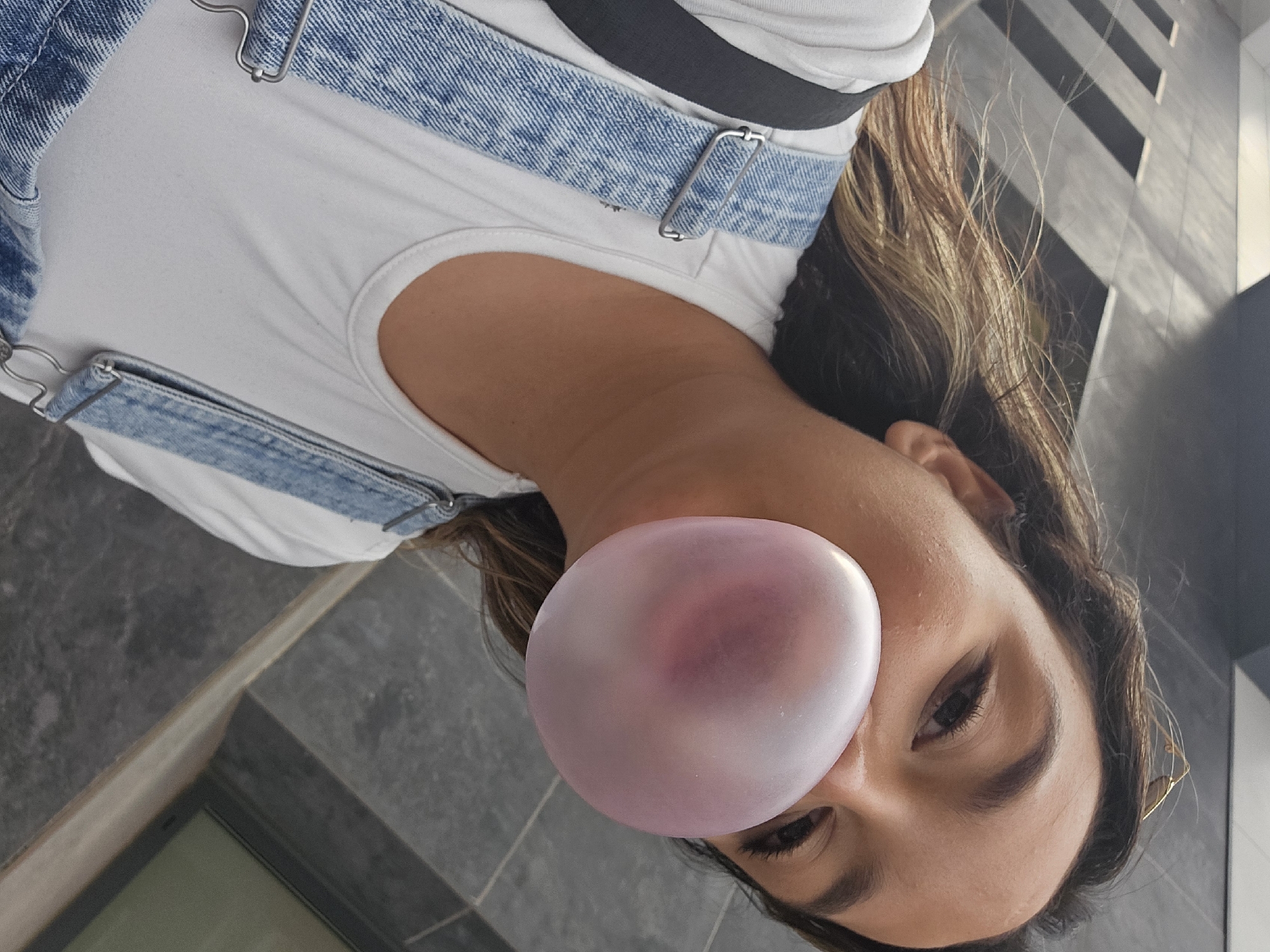 In a medium-close-up, low-angle shot, a young woman with long, wavy brown hair is captured from the chest up, blowing a large pink bubble gum bubble. She is wearing a white tank top and blue denim overalls with metal clasps. Her eyes are focused on the camera, and her mouth is open, forming the bubble. The background appears to be an urban environment, with textured grey walls and streaks of sunlight and shadow suggesting an outdoor setting. The scene is bright, with the sunlight highlighting the bubble and the woman's features. The overall impression is one of a casual, playful moment captured outdoors. The location of Castelló de la Plana, Spain is not identifiable from the visual information in the image.