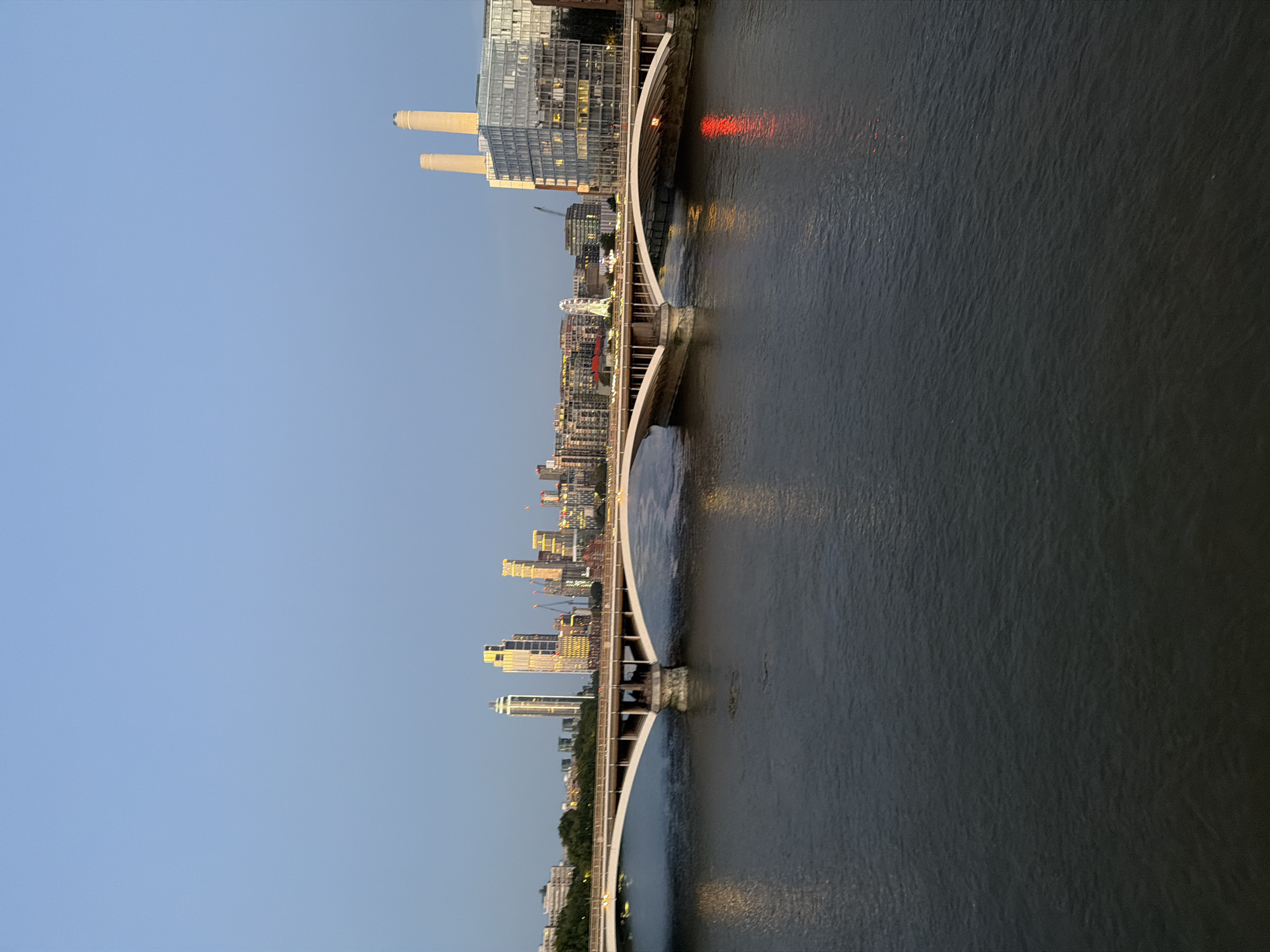 The image shows a view of the River Thames in London, United Kingdom, with Battersea Power Station visible in the background. A multi-lane road bridge spans the river, with traffic and streetlights visible on its surface. The water of the Thames is dark and reflects the lights from the city. In the distance, a cluster of modern buildings, including some tall skyscrapers, lines the riverbank. Battersea Power Station, a landmark building with its distinctive four chimneys, is prominently featured, with a modern glass-fronted building adjacent to it. The sky is clear and a pale blue, suggesting it is either early morning or late evening. There are no people or vehicles clearly discernible in the image. The scene depicts an urban landscape at dusk or dawn, highlighting the contrast between historic and contemporary architecture.