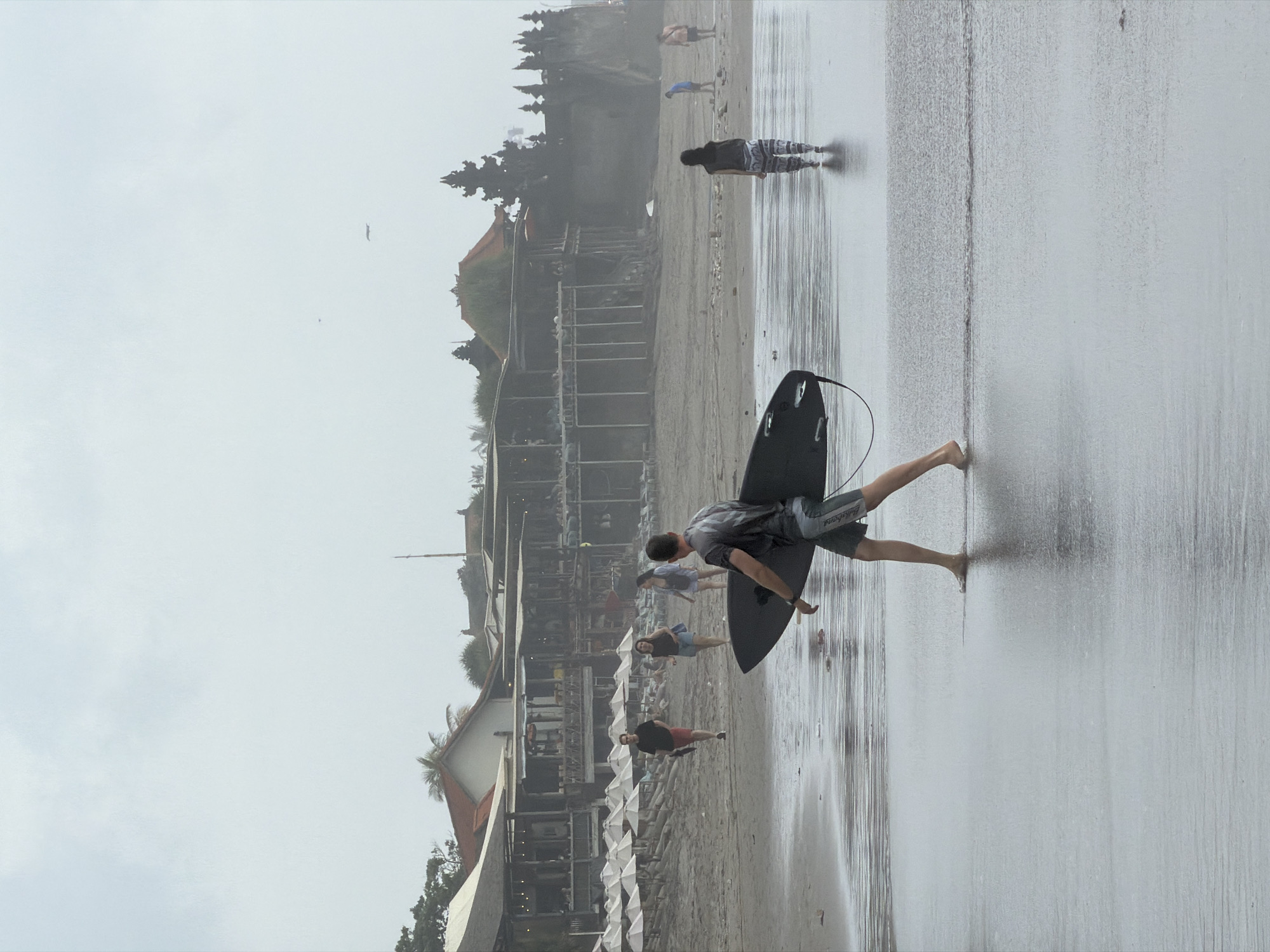 In North Kuta, Indonesia, on a cloudy day, a person walks along the wet sand at the water's edge carrying a black surfboard. The person is wearing a grey t-shirt with a pattern and grey shorts. In the background, several other people are walking on the beach, and a row of beach umbrellas is visible. Beyond the beach, there are buildings with red roofs, including what appears to be a restaurant or resort, and a temple structure on a cliff. The sky is overcast, and the light is diffused, creating a slightly hazy atmosphere. The ocean is calm with small waves washing ashore.