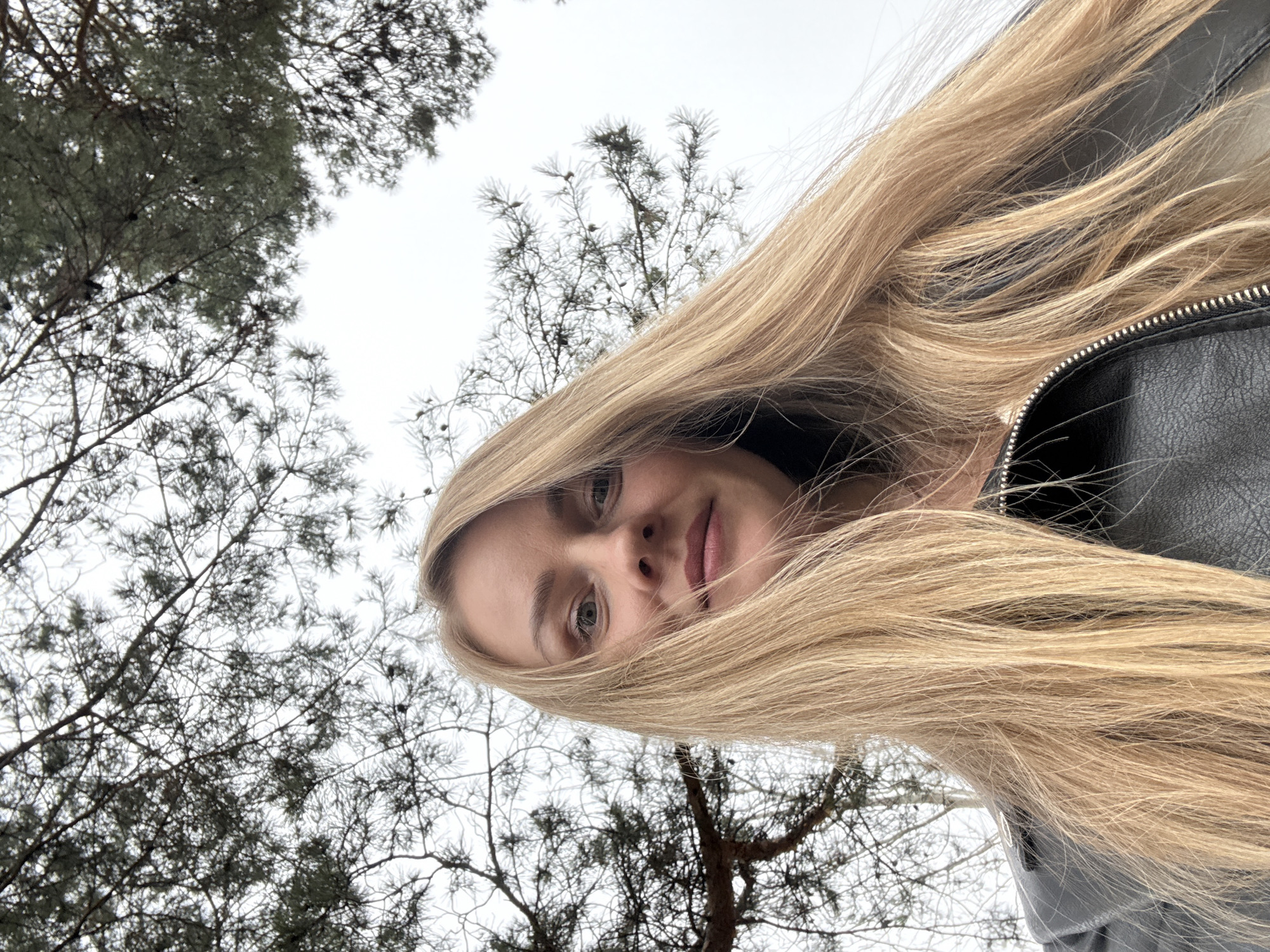 A blonde woman wearing a black leather jacket poses outdoors in Chaikovsky, Russia. The photo is taken from a low angle, looking up at the woman. Bare trees and an overcast sky are visible in the background.