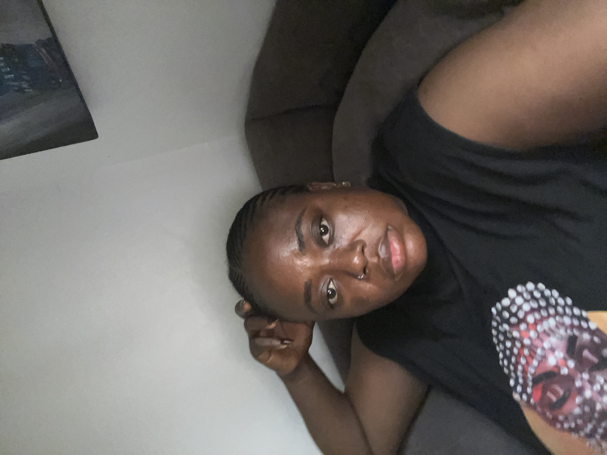 A dark-skinned woman with braided hair and a nose ring is captured in a close-up, frontal shot. She is reclining on a dark brown couch, with her left arm bent and her hand resting on her head. The woman is wearing a sleeveless black top. Her right arm is partially visible, holding a cylindrical object adorned with a mosaic pattern of red, white, and black elements. The background consists of a light-colored wall, with a portion of a framed artwork visible in the upper right corner. The overall lighting is dim, with shadows accentuating the contours of her face. The environment appears to be indoors, likely a residential setting in Abuja, Nigeria.