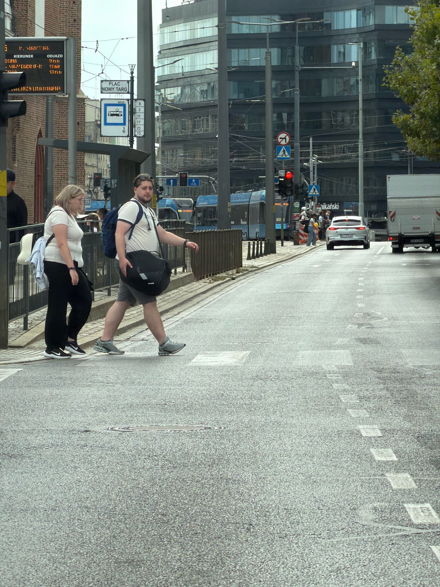 Two individuals are crossing a street in Wrocław, Poland, at a pedestrian crossing. The person on the left is a woman wearing glasses, a white t-shirt, and black pants, carrying a light-colored garment over her left shoulder. The person on the right is a man wearing a white t-shirt, grey shorts, and a dark backpack, carrying a large black duffel bag in his right hand. They are walking from left to right across the asphalt road, which has dashed white lines marking the pedestrian crossing.
In the background, several elements indicate an urban setting. To the left, a brick building with a digital display board is visible, showing information in Polish. A traffic light, currently red, is mounted on a pole. Further back, a modern glass-fronted building with multiple floors stands tall. Several blue trams are parked or moving along a tram line in the middle ground. To the far right, a white truck is parked. Various street signs, including one indicating "PLAC NOWY TARG" (New Market Square) and tram route numbers, are present. The sky is overcast.