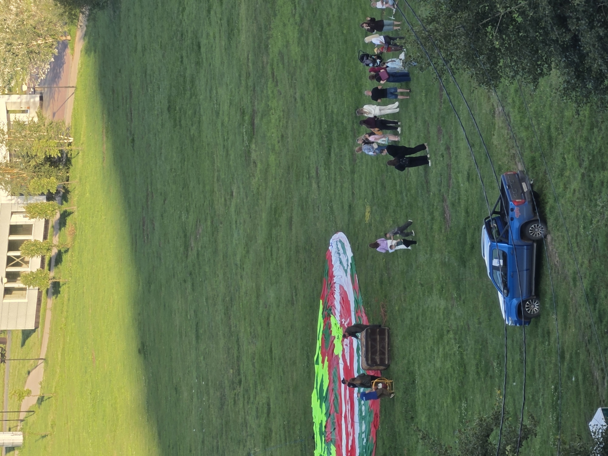 The image, taken from an elevated perspective in Nizhny Novgorod, Russia, depicts a group of people gathered on a grassy field. In the foreground, a blue pickup truck is parked on the grass. Further back, a large, deflated hot air balloon with a red, green, and white pattern is spread across the ground. Several individuals are interacting around the balloon and what appears to be the balloon's basket. To the right, a larger group of people is standing and observing. The environment includes a paved walkway and a building in the background, suggesting an urban or suburban park setting. The lighting indicates it is likely late afternoon or early evening.