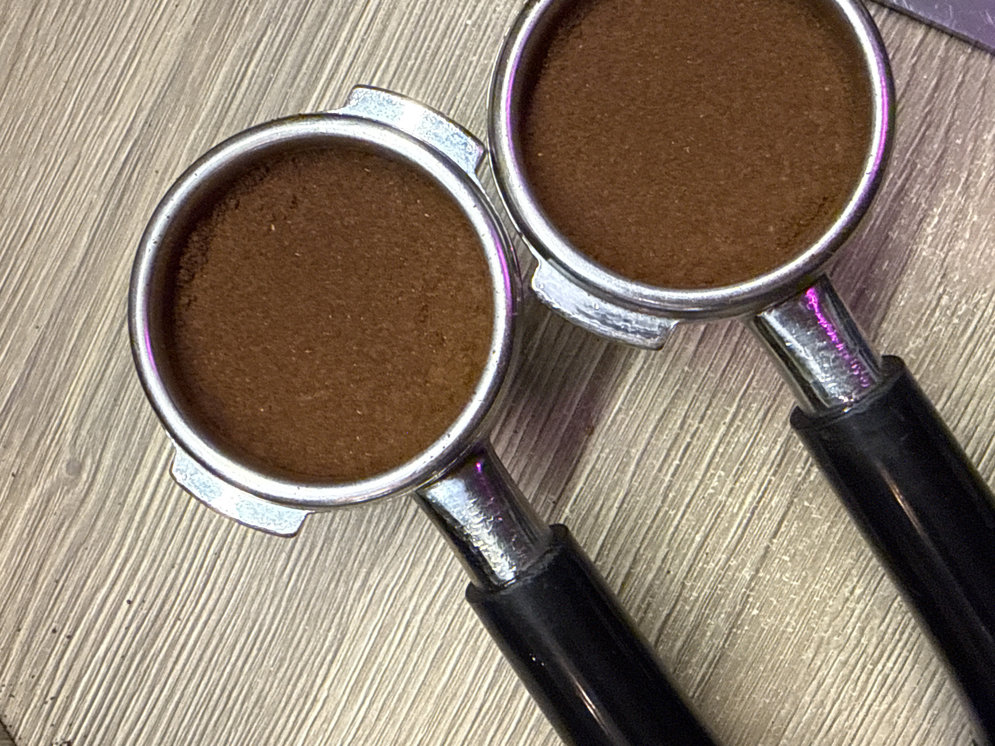The image depicts two espresso portafilters filled with ground coffee. They are positioned side-by-side on a surface with a wood-grain pattern. The portafilters are stainless steel with black handles. The ground coffee inside each is evenly distributed and appears freshly tamped. There is no context to connect this image to the city of Dolgoprudny, Russia.