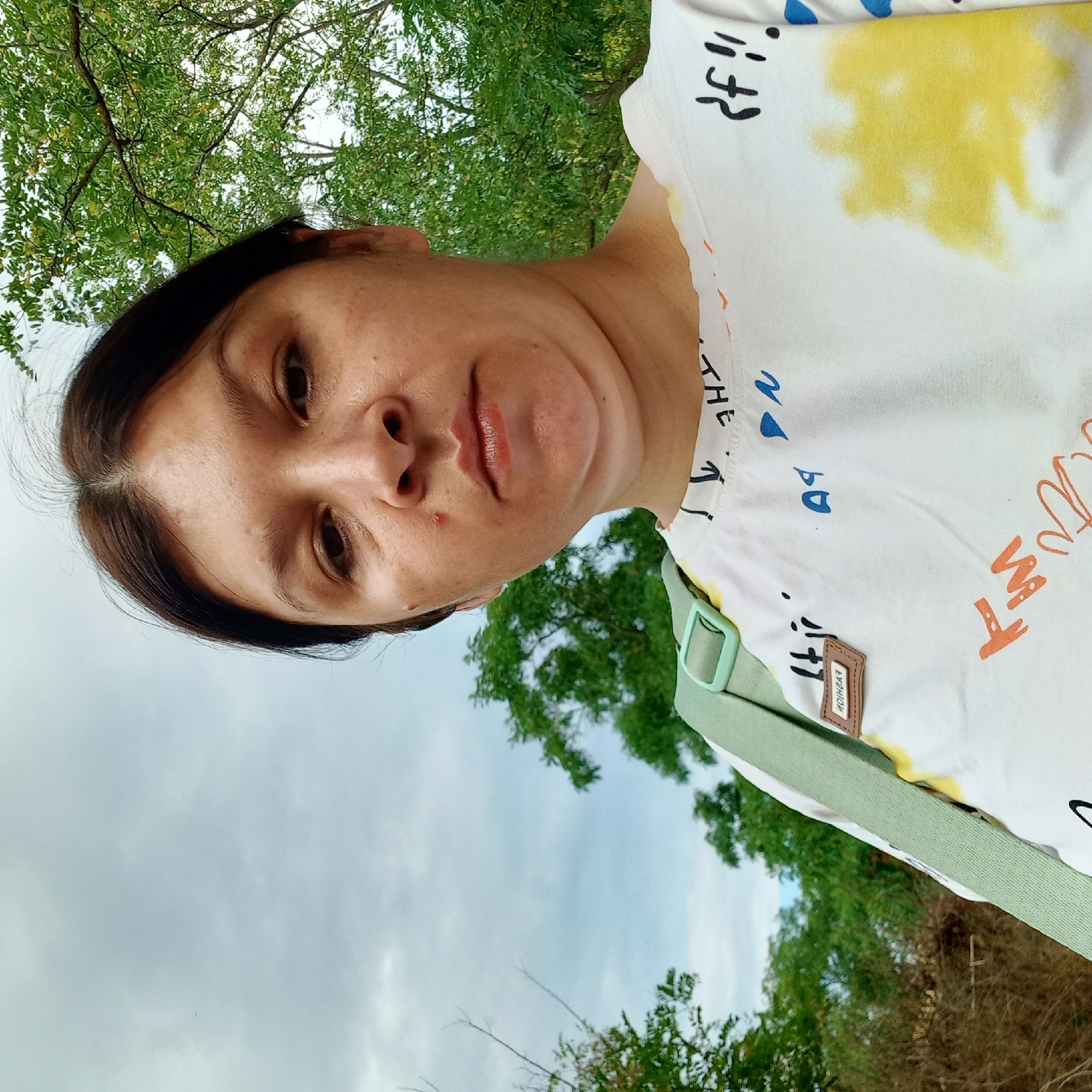 The image shows a close-up, low-angle selfie of a woman outdoors. She has brown hair and is looking directly at the camera with a neutral expression. She is wearing a white t-shirt with colorful abstract designs and text. A light green strap from a bag is visible across her shoulder. The background consists of green trees and a cloudy sky, suggesting a natural or park-like setting. The location in the city of Klyntsi, Ukraine, is not visually indicated in the image.