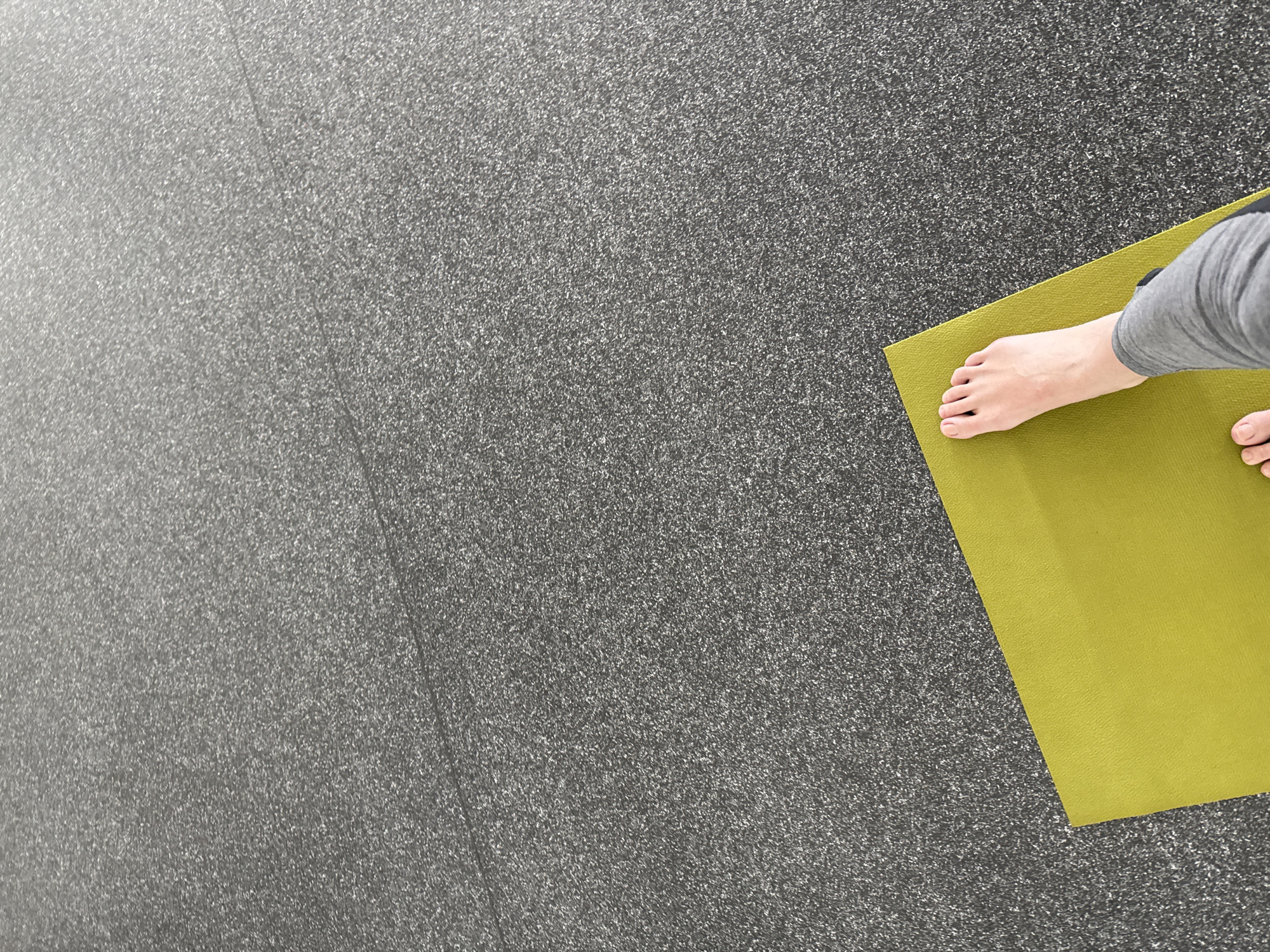 A high-angle, medium shot presents a person standing on a yellow-green yoga mat against a speckled gray floor. The individual's bare feet and lower legs, clad in gray, are visible on the right side of the frame, positioned on the mat. The yoga mat itself is a solid, muted yellow-green color, occupying a small area in the lower right corner. The background consists of a uniformly speckled, dark gray floor, its texture resembling a fine grain. The floor extends across the majority of the image, creating a neutral backdrop. The scene is evenly lit, contributing to the distinct separation of the objects.