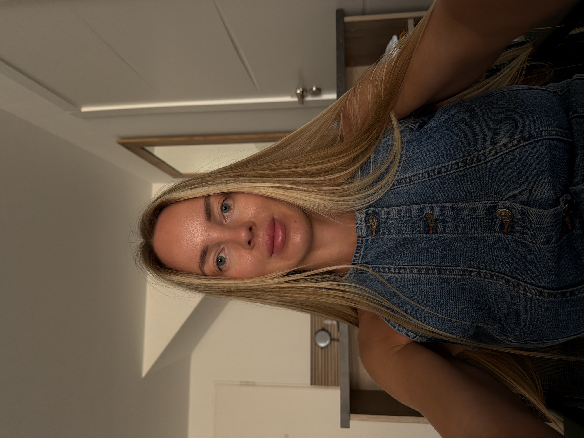 The image is a selfie of a blonde woman with blue eyes, wearing a denim top. She is seated indoors, in what appears to be a room with a light-colored wall and a door with a silver handle in the background. To the left of the woman, there is a piece of furniture with a mirror and a round object on it. The lighting suggests it might be evening or early morning. The woman's hair is long and blonde, framing her face. Her skin is tanned. The background environment does not provide specific clues to confirm the location as Finestrat, Spain.