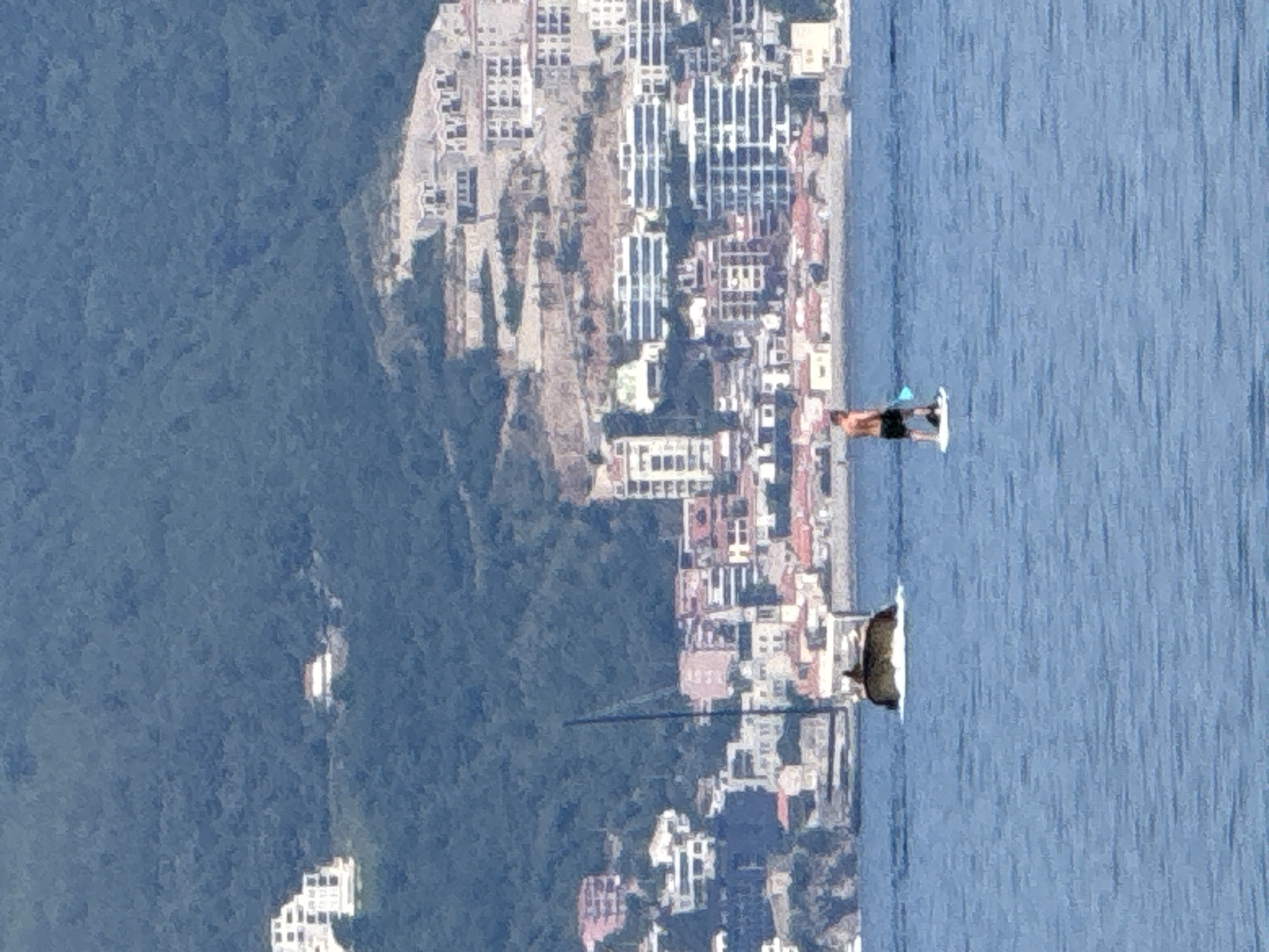 The image shows a person stand-up paddleboarding on a body of water. The person is in the middle of the frame, facing away from the camera, and appears to be wearing black shorts and a dark top. They are holding a paddle. Behind the paddleboarder, a town is visible built into the side of a lush green hill, overlooking the water. The buildings are predominantly white and light-colored, with red-tiled roofs. In the distance, more buildings are visible, suggesting a densely populated area. The water is a calm, deep blue with subtle ripple patterns. The scene is bathed in natural light, suggesting daytime. The overall impression is one of leisure and activity in a scenic coastal environment. The location is Boreti, Montenegro.