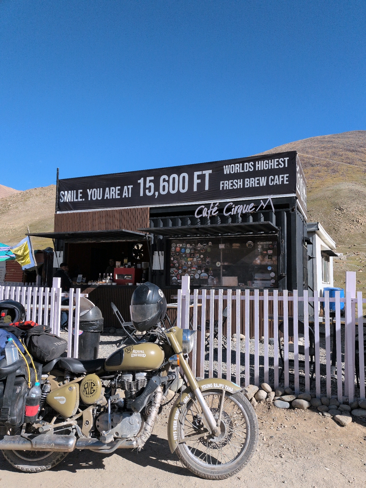 The image depicts "Cafe Cirque," advertised as the "Worlds Highest Fresh Brew Cafe" at 15,600 feet, located in Khardung La, Ladakh, India. The cafe is a converted shipping container situated on a mountainside under a clear blue sky. A prominent black banner above the cafe reads "SMILE. YOU ARE AT 15,600 FT WORLDS HIGHEST FRESH BREW CAFE". The cafe's front features a serving window decorated with numerous stickers, and two individuals are visible inside, presumably staff.
In the foreground, a khaki-colored Royal Enfield motorcycle is parked. The motorcycle is equipped with saddlebags and other luggage, suggesting it is used for travel. Two helmets are placed on the motorcycle and nearby. A white picket fence separates the cafe area from the motorcycle and the viewer's perspective. The ground is composed of gravel and dirt, with a pile of rounded stones next to the fence. The background consists of a barren, rocky mountainside.