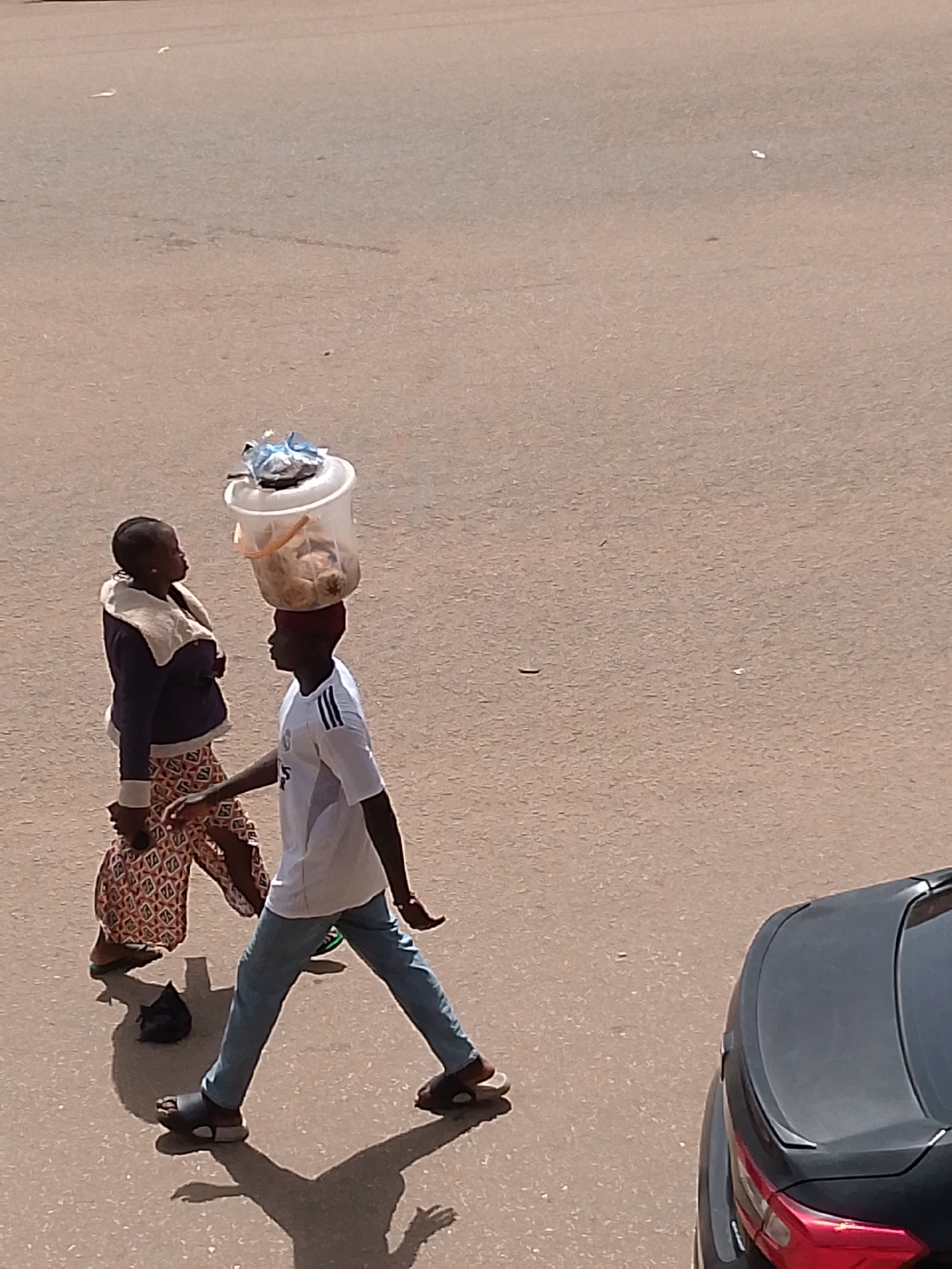 In Bukuru, Nigeria, a man walks carrying a large clear plastic bucket filled with food items on his head. He wears a white t-shirt with black stripes on the sleeves, blue jeans, and dark sandals. Beside him, a woman walks with her left hand on her hip. She is wearing a dark jacket with a fluffy collar and a patterned skirt. A black bag lies on the ground near her feet. In the background, the top of a black car is visible. The scene takes place on a paved road with the sun casting long shadows.