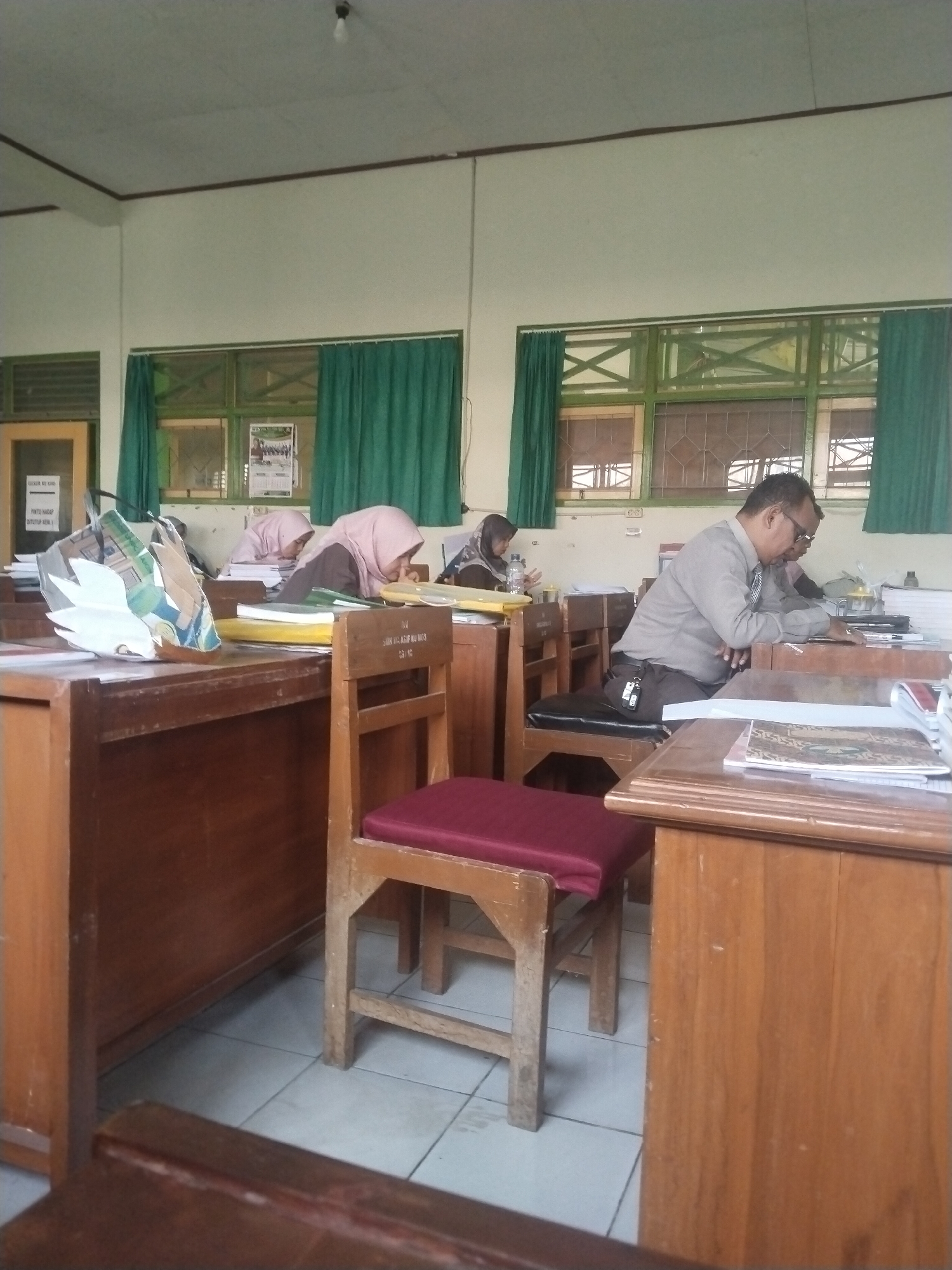 The image depicts a classroom scene. Two female students, wearing headscarves, are seated at their desks, appearing to be engaged in their work. One student is looking down at her desk, while the other is partially visible in the background. A male individual, wearing glasses and a light-colored shirt, is also seated at a desk, facing away from the camera and seemingly focused on paperwork. The room features wooden desks and chairs, with green curtains covering the windows. The overall environment suggests an educational setting, possibly a school in Pakulaut, Indonesia, given the typical classroom setup and attire of the individuals. There is a bag on one of the desks in the foreground. The lighting appears to be from overhead, with a single bulb visible on the ceiling.