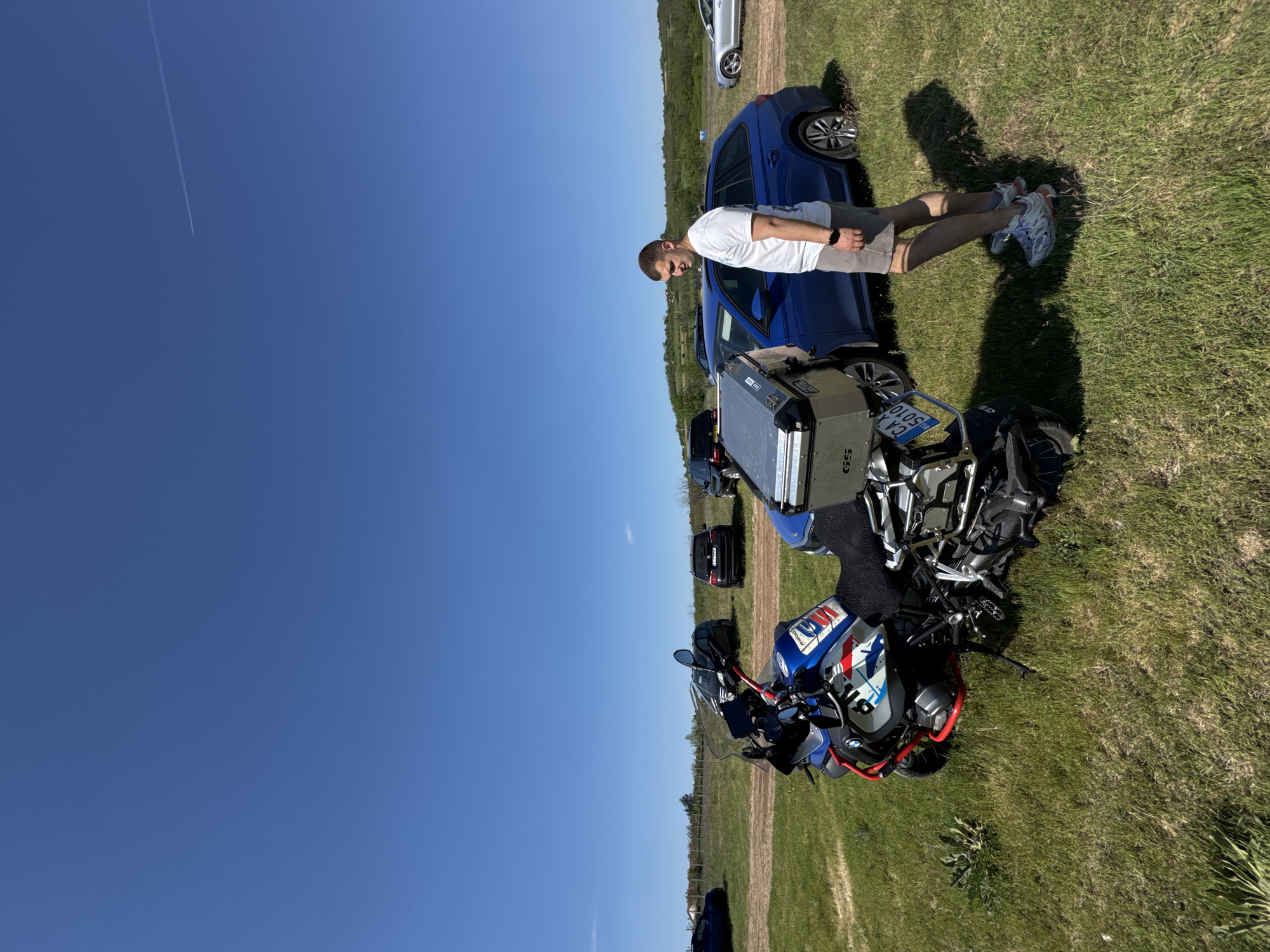 A sunny outdoor scene in Lukovit, Bulgaria shows a man in a white t-shirt, tan shorts, and sneakers standing near a blue BMW motorcycle with saddlebags. A blue Volkswagen car is parked nearby. The ground is grassy. In the distance, several other cars are visible along with a tree line. The sky is clear blue with a contrail from an aircraft.