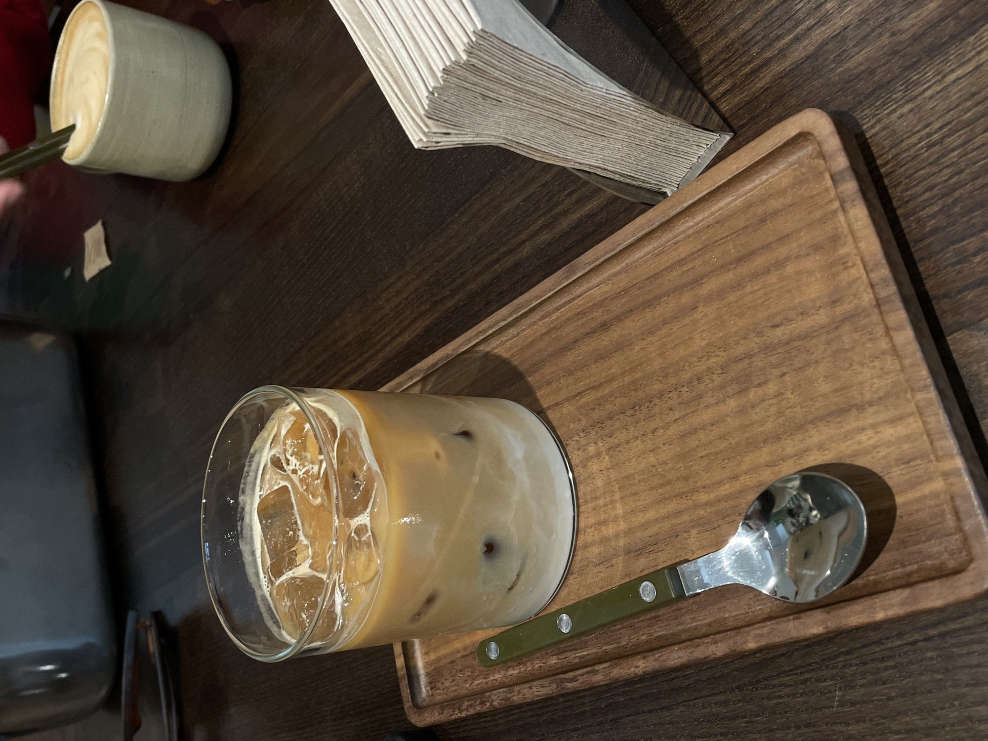 On a dark wooden table in Milan, Italy, sits a wooden tray containing an iced coffee in a glass, a spoon with a green handle, and a stack of folded napkins. To the left, there is a ceramic cup containing coffee or a latte. The scene is bathed in natural light. No people are visible.