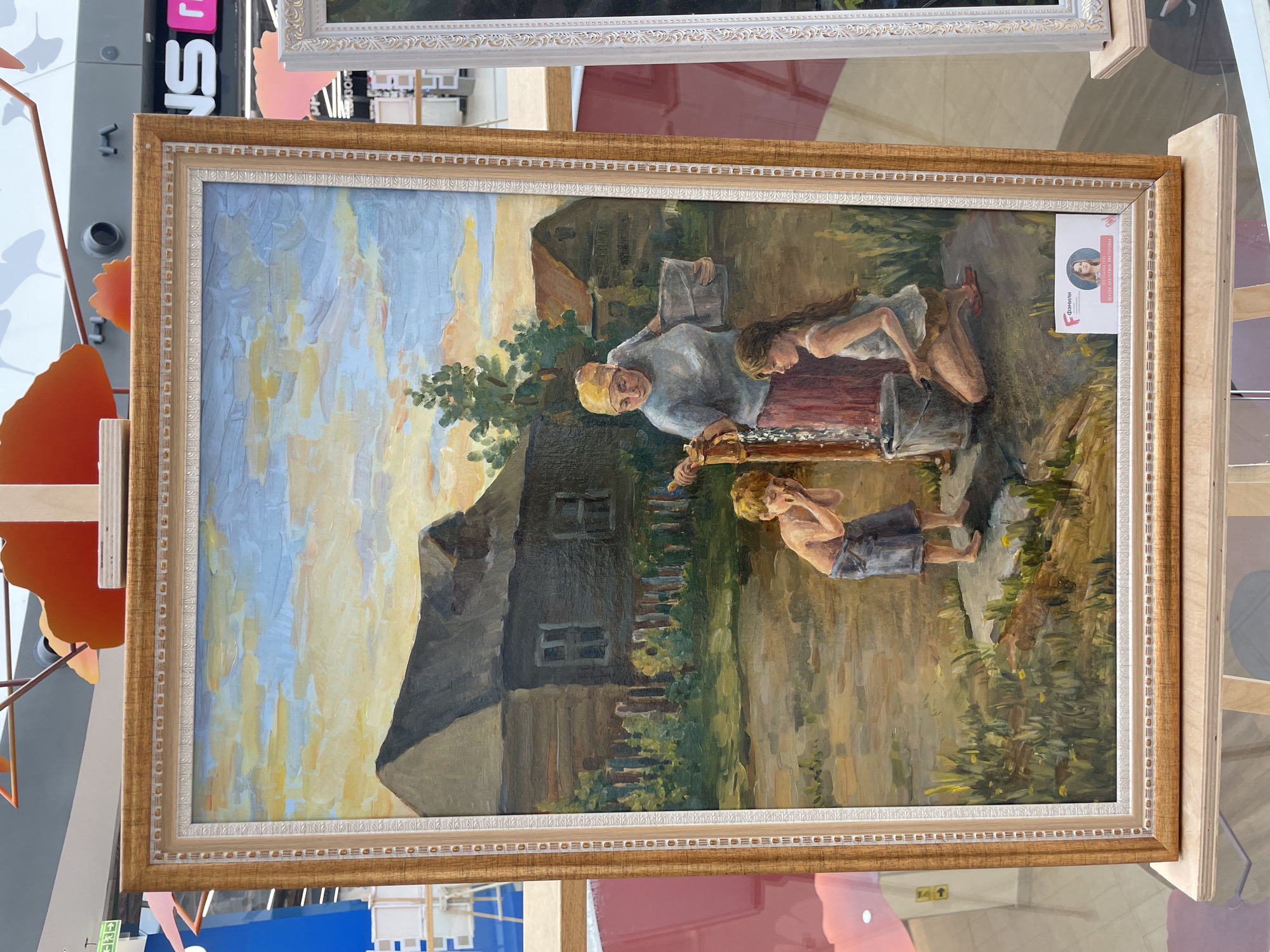 This is a painting depicting a rustic scene, likely in a rural setting in Perm, Russia. The painting features three individuals: an older person, possibly a grandmother, wearing a headscarf and a blue tunic, and two younger people, a girl with long hair and a boy wearing shorts. The older person appears to be engaged in a task, possibly washing something in a bucket or trough. The girl is leaning in, her attention focused on the activity, while the boy stands nearby, looking towards the water.
The scene is set outdoors, with a dark, weathered house in the background, suggesting a humble dwelling. A wooden fence and lush green foliage add to the rural atmosphere. The sky is rendered with a blend of blue and yellow hues, indicating it might be late afternoon or early evening, possibly during a sunny day. The overall impression is one of domestic activity and a sense of community within a simple, rural environment.
A small white card is attached to the right side of the painting's frame. It contains some text and a circular image of a woman with blonde hair. The text is in Cyrillic, and while not fully legible, the word "Фокил" is visible. This card likely provides information about the artwork or the artist.