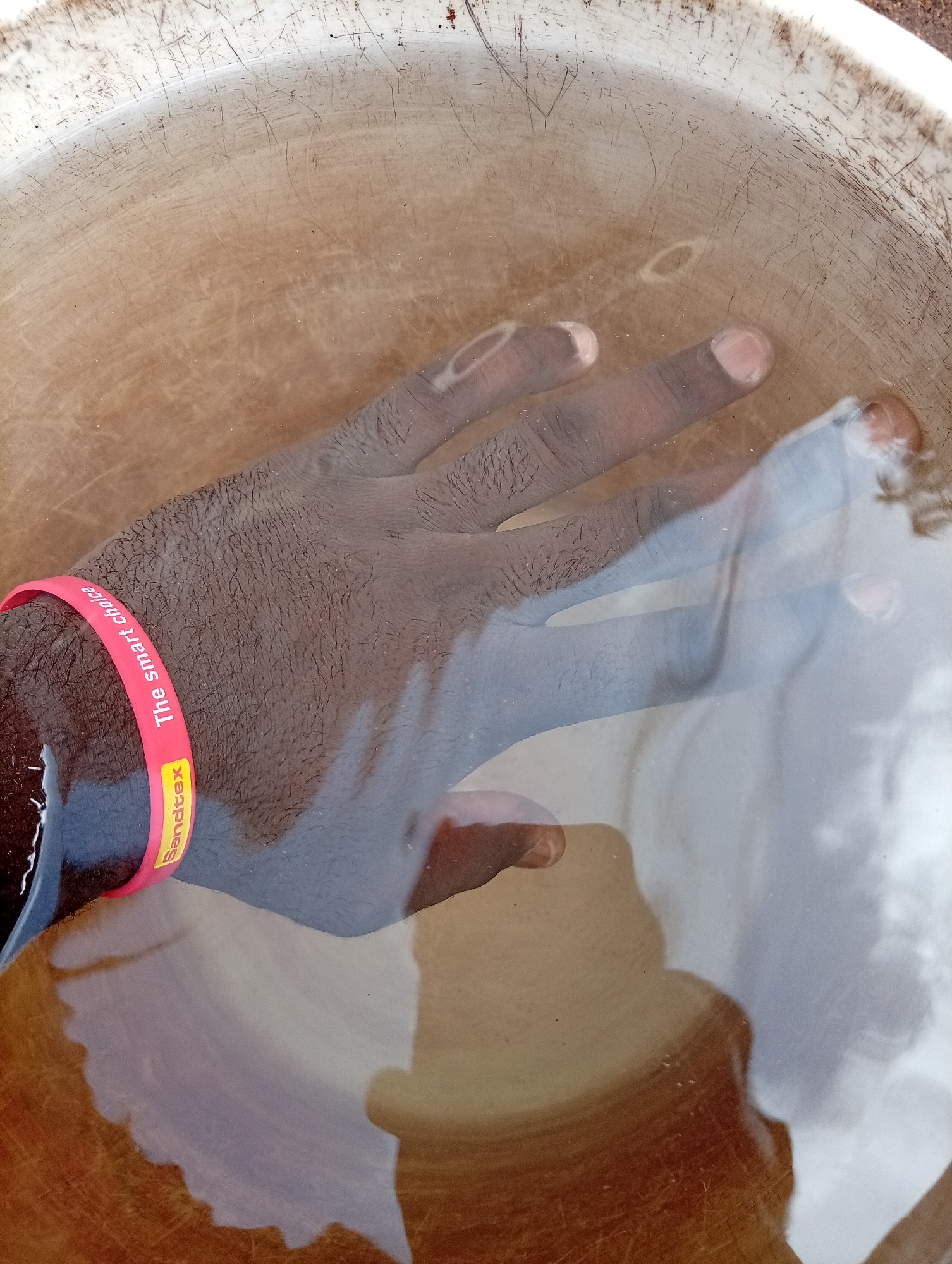 A dark-skinned human hand, wearing a red wristband with the text "Sanditex The smart choice", is submerged in a liquid within a light-colored, possibly plastic, basin. The liquid appears to be water, possibly with some discoloration or sediment. The inside surface of the basin shows scratches and marks. No other specific information suggesting the city of Bukuru, Nigeria, is visible.