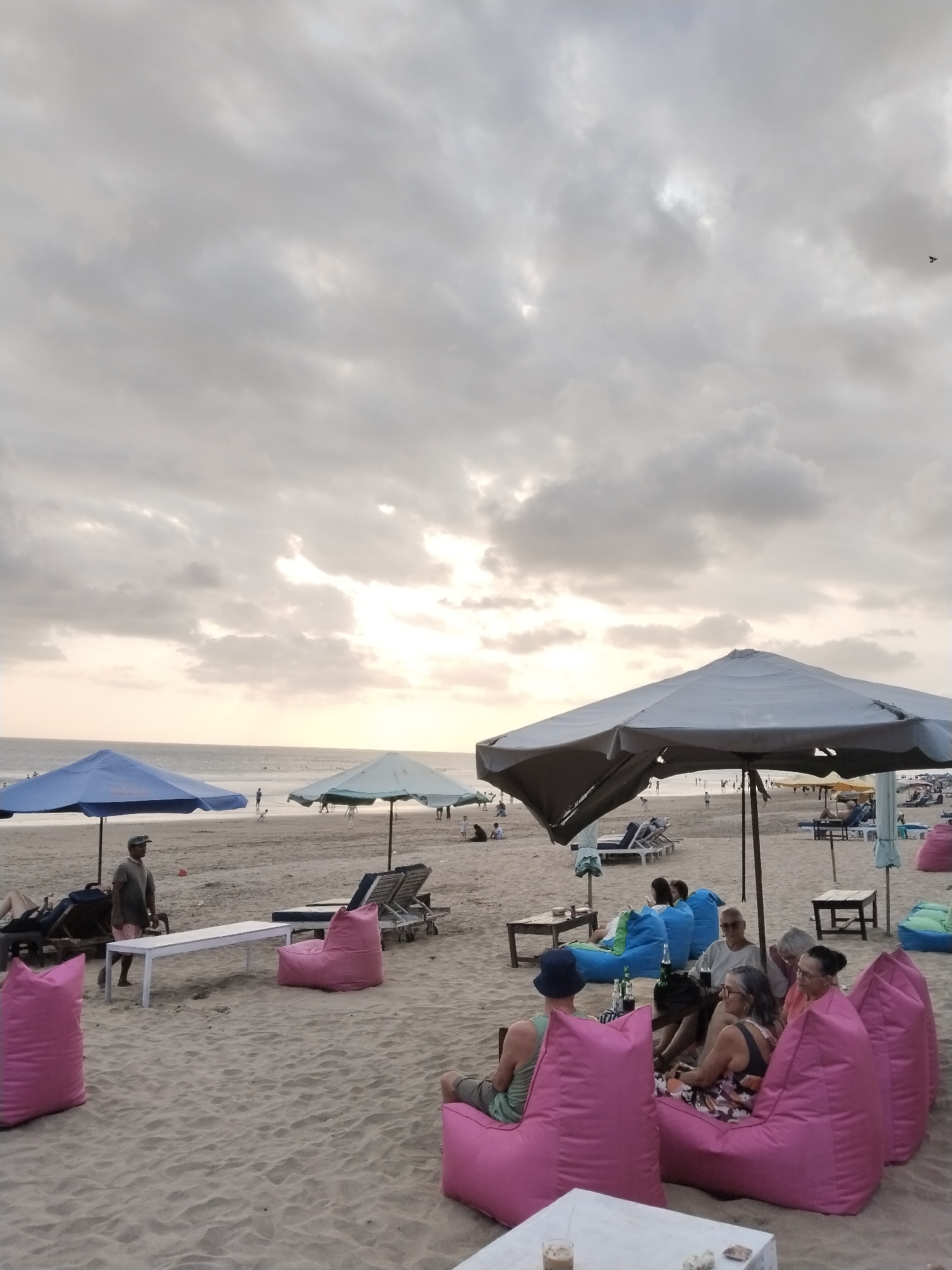The image depicts a cloudy sky over Kuta Beach in Bali, Indonesia, during the late afternoon or early evening. The sand is light brown, and the sea is visible in the distance. Several beach umbrellas, predominantly blue and gray, are set up on the sand. In the foreground, there are bright pink beanbag chairs, some occupied by people. A group of individuals, including men and women, are seated on pink and blue beanbag chairs, some interacting with each other, while others appear to be looking out towards the sea or engaged with their drinks. In the mid-ground, a man in a dark t-shirt and shorts stands near a white table and lounge chairs. Further back, other people are visible on the beach, some sitting under umbrellas, others walking or playing. The overall atmosphere suggests a relaxed beachside setting.