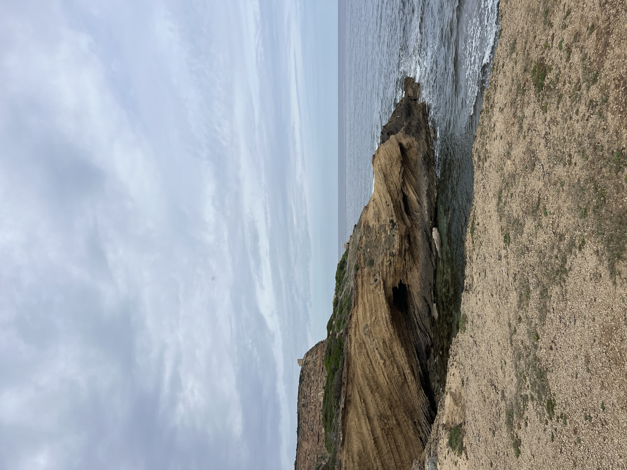 The image depicts a rocky coastline under an overcast sky. To the right is a gravelly cliff edge covered with sparse vegetation. In the middle is a large rock formation with layered geological strata, with a small cave-like opening at its base. The sea borders the rocks, with small waves crashing against them. In the distance, the sea meets the sky in a seamless horizon line. A tower is visible atop a cliff on the left side. The scene is located in San Vero Milis, Italy.