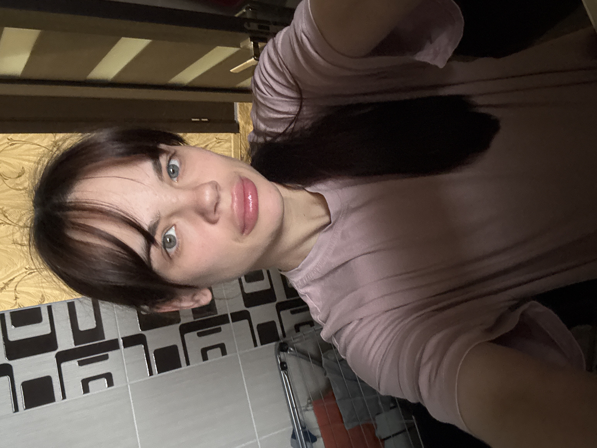 A selfie of a woman with light skin, blue-grey eyes, and dark brown hair pulled back in a low ponytail. She is wearing a light pink t-shirt. The background consists of a black and white tiled wall, a metal towel rack with folded towels, and a wooden ceiling. The location cannot be determined to be Rostov-on-Don, Russia from the provided image.