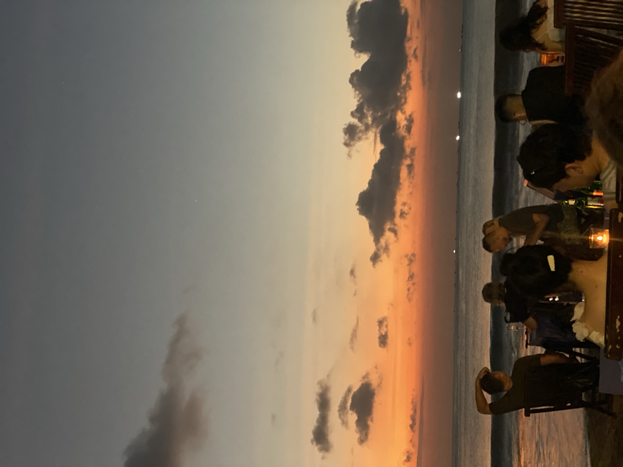At dusk in Kuta Selatan, Indonesia, a group of people are seated at tables on a beach facing the ocean. The sky is painted with hues of orange, pink, and gray as the sun sets. The water is calm with gentle waves lapping the shore. Some of the individuals are facing the sea, while others are engaged in conversation. There are tables set up with what appear to be drinks and possibly food. In the distance, there are lights visible on the water, suggesting boats or offshore structures. The overall atmosphere is relaxed and serene, with the natural beauty of the sunset setting the scene.