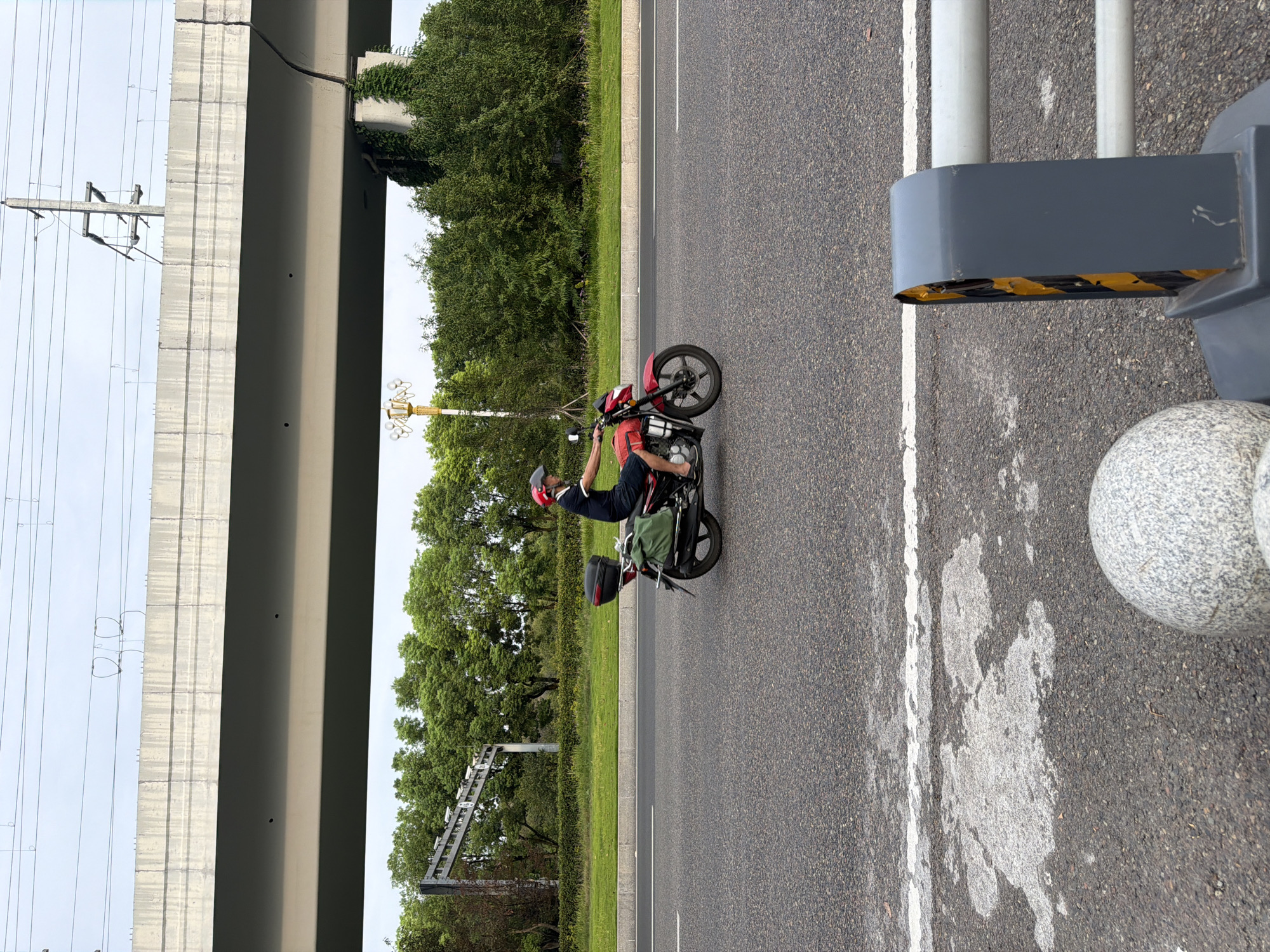 A male rider wearing a red helmet and a dark blue shirt rides a red motorcycle on a paved road in Jiangjin District, Chongqing, China. The motorcycle has a black storage box on the rear and a green bag strapped to the side. The rider is wearing dark shorts and appears to be barefoot. The road is bordered by a grassy median and a line of trees. An elevated concrete structure, likely a highway overpass, spans across the background, with power lines and utility poles visible above it. In the foreground, a concrete bollard and a gray barrier with black and yellow hazard markings are present. The sky is overcast.